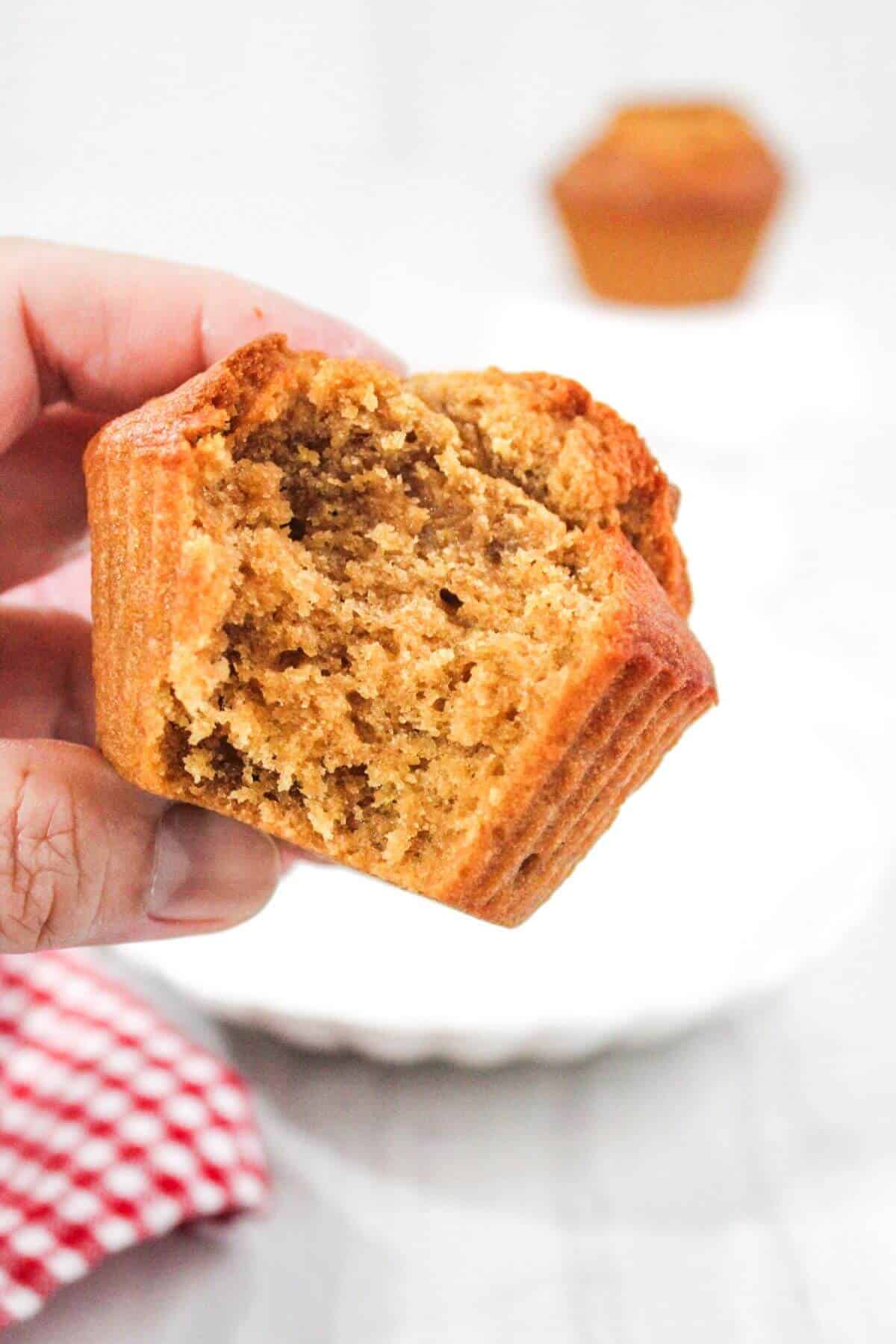 A hand holds a partially eaten muffin with a textured, crumbly interior. A second muffin is blurred in the background. A red and white checkered cloth is partially visible in the bottom left corner.