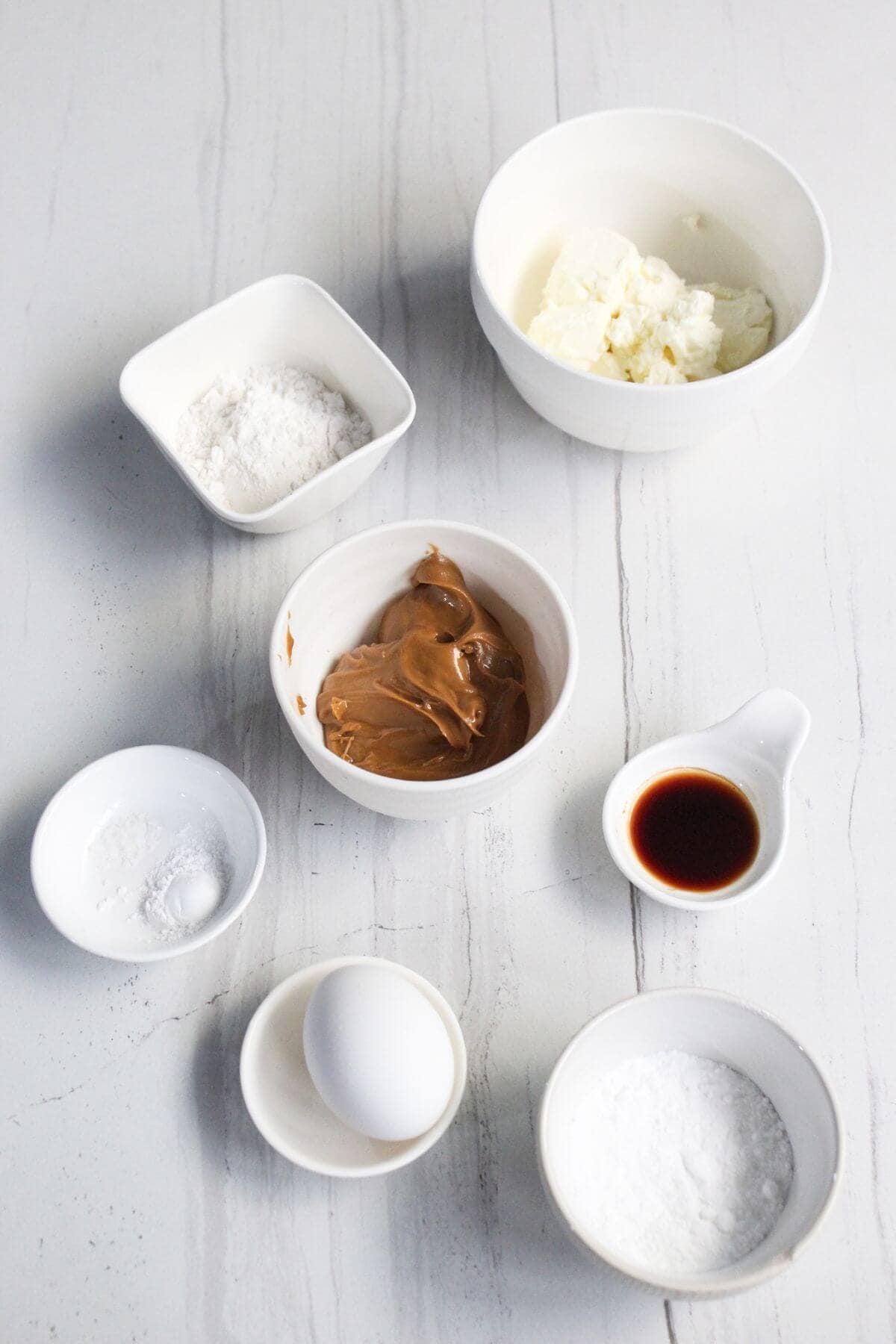 Various baking ingredients are arranged in white bowls on a white surface, including flour, cream cheese, peanut butter, vanilla extract, baking soda, an egg, salt, and powdered sugar.