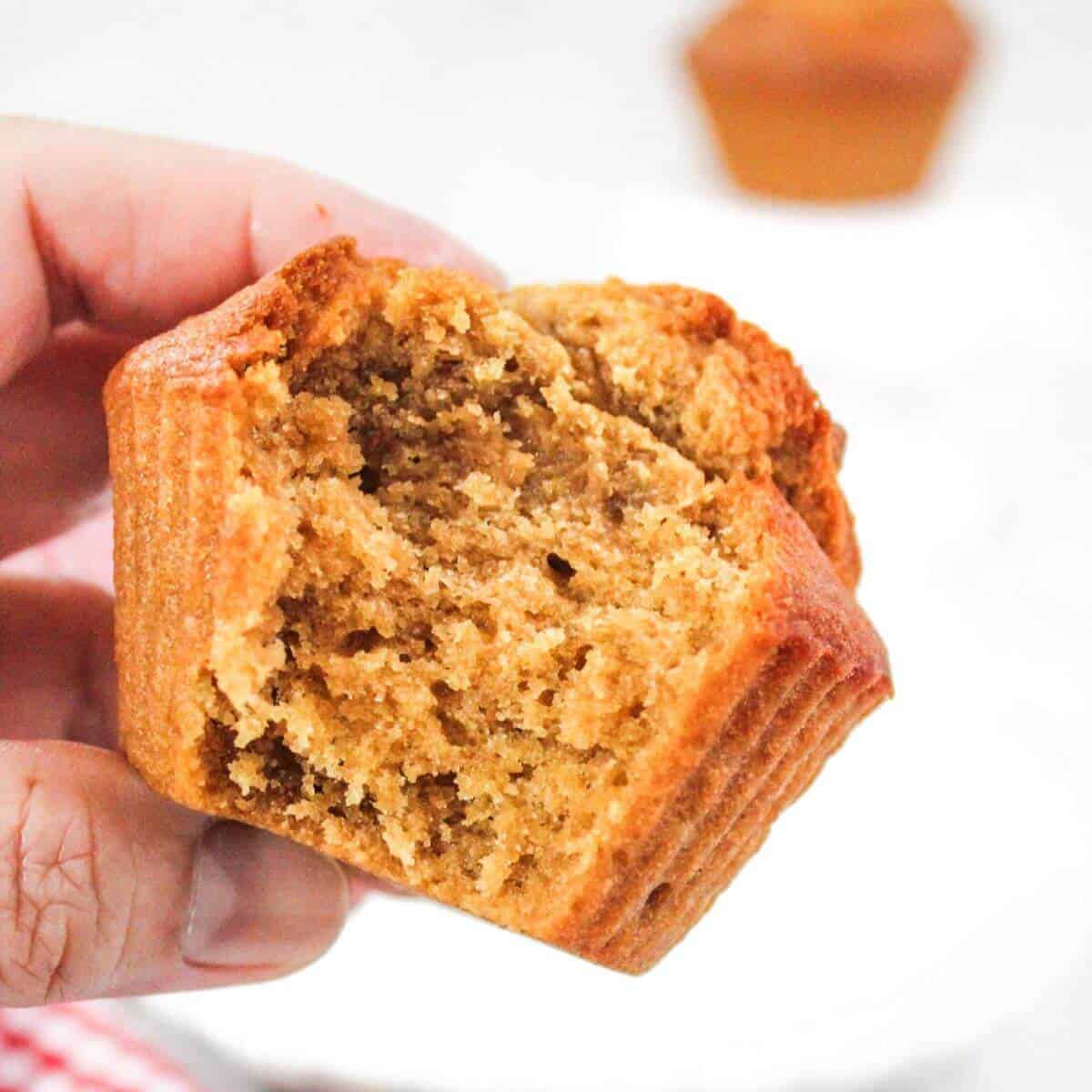 A close-up of a hand holding a bitten-into, golden-brown muffin with a soft, crumbly texture. Another muffin is blurred in the background.