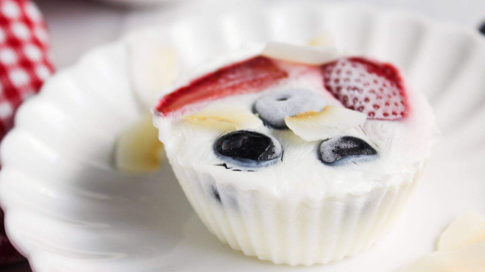 A white cupcake liner holds a yogurt dessert with blueberries, strawberry slices, and almond slivers on top, presented on a white plate.