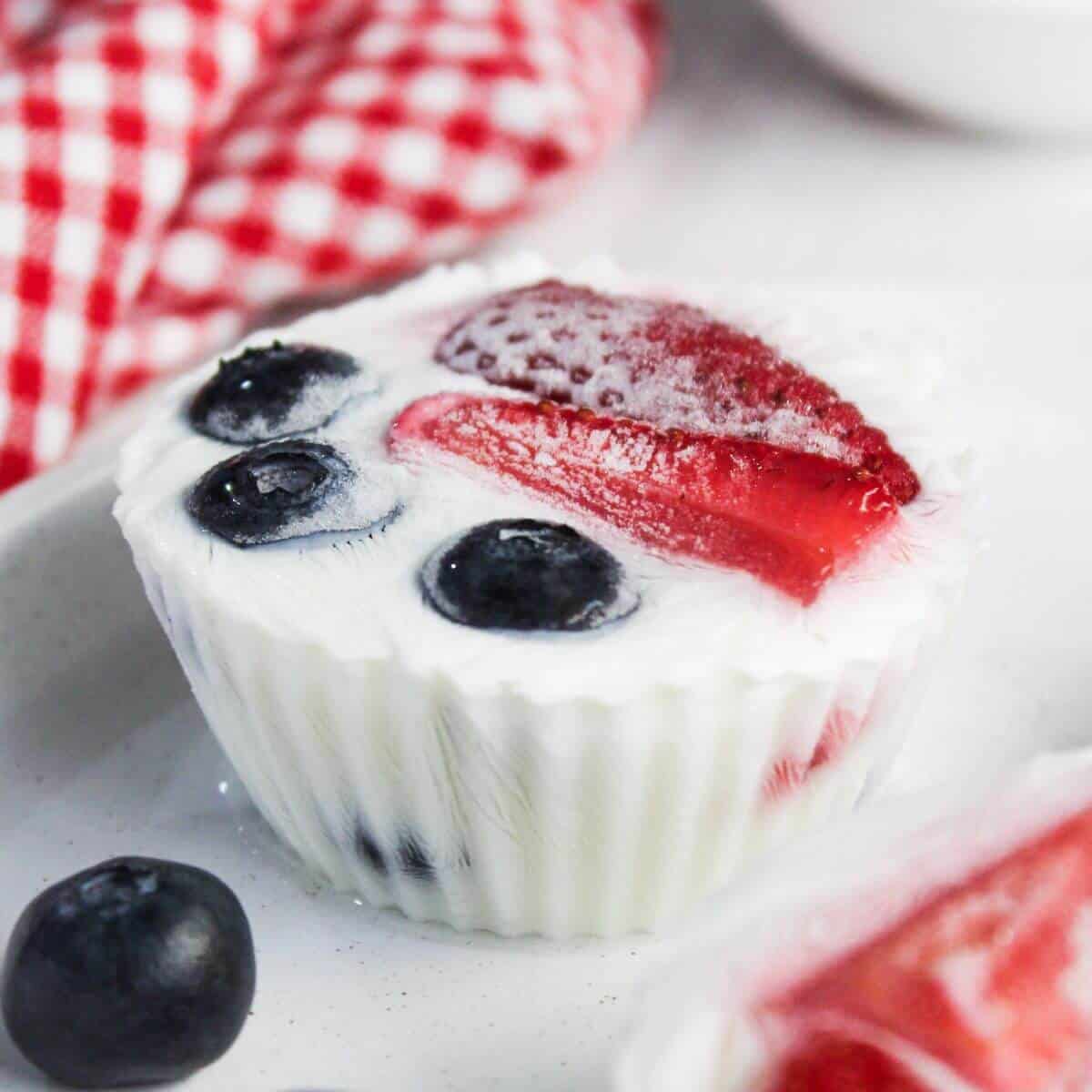A white yogurt cup topped with sliced strawberries and whole blueberries, with a red and white checkered cloth in the background.