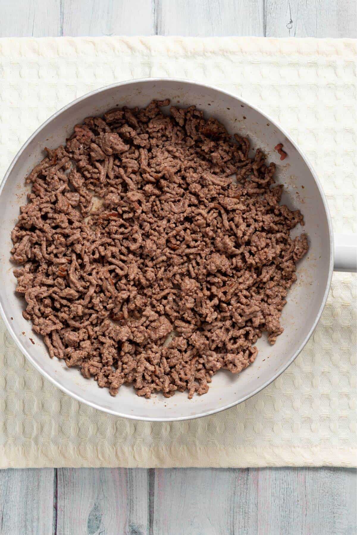 A white frying pan filled with cooked ground beef sits on a light-colored, textured cloth on a wooden surface.