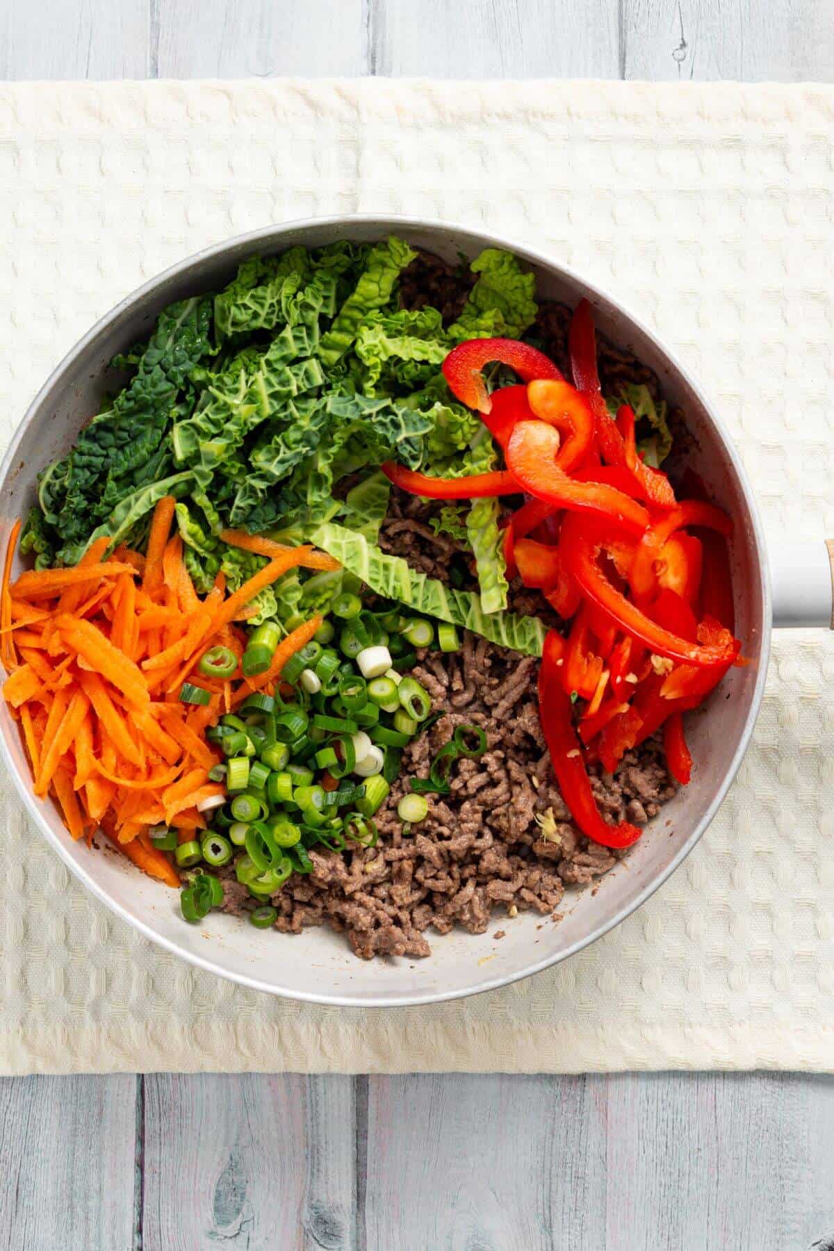 A pan contains sautéed ground meat, shredded green cabbage, sliced red bell peppers, chopped green onions, and julienned carrots on a white textured surface.