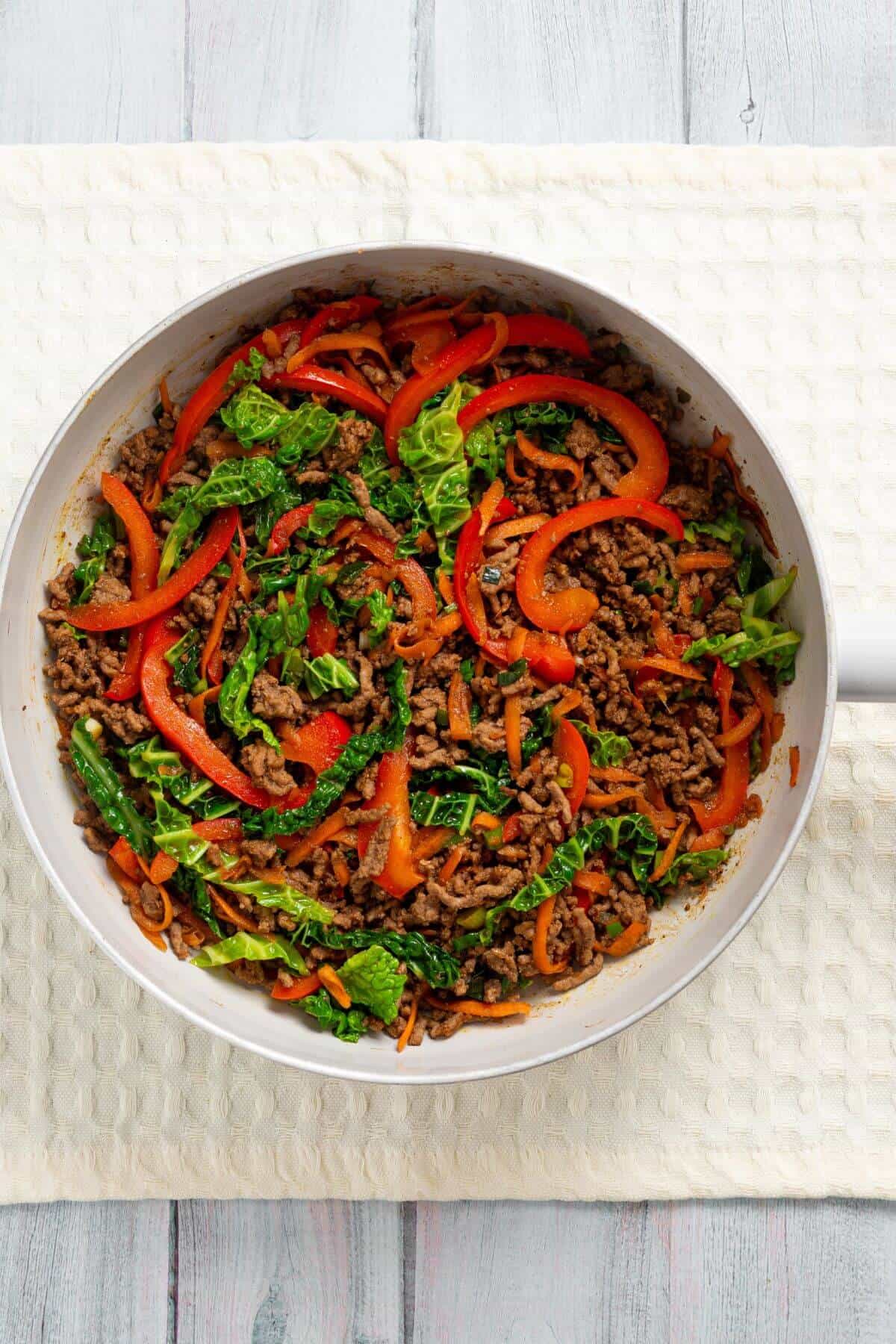 A skillet filled with cooked ground beef, sliced red bell peppers, shredded carrots, and chopped leafy greens sitting on a white surface.