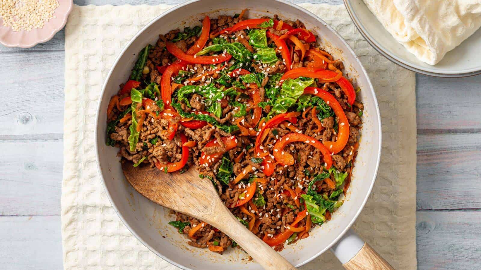 A skillet filled with a colorful stir-fry of ground meat, bell peppers, leafy greens, and sesame seeds, with a wooden spoon resting in it. Plates and a stack of flatbreads are nearby.