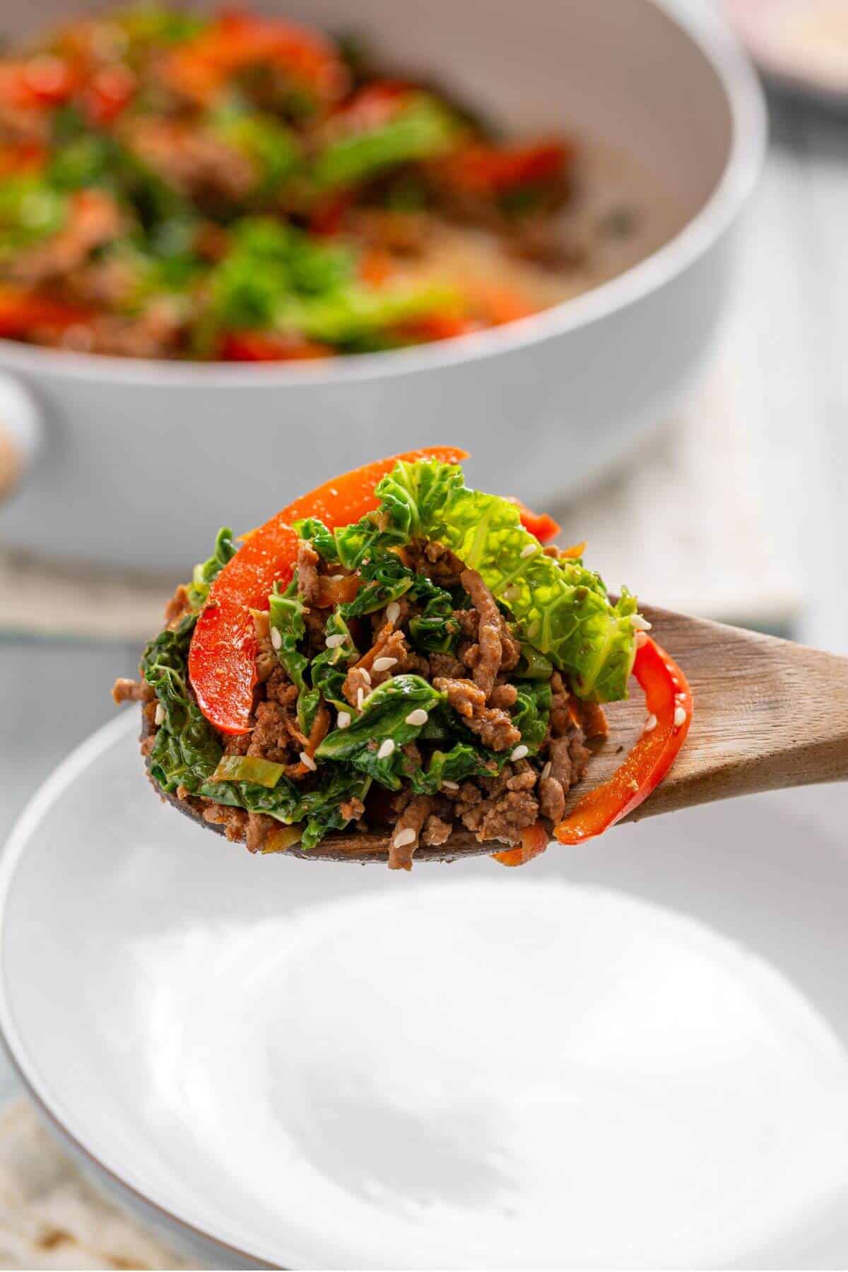 A wooden spoon holds a portion of a stir-fry dish with ground meat, red bell peppers, and greens over a white bowl. A pan with the same dish is in the background.