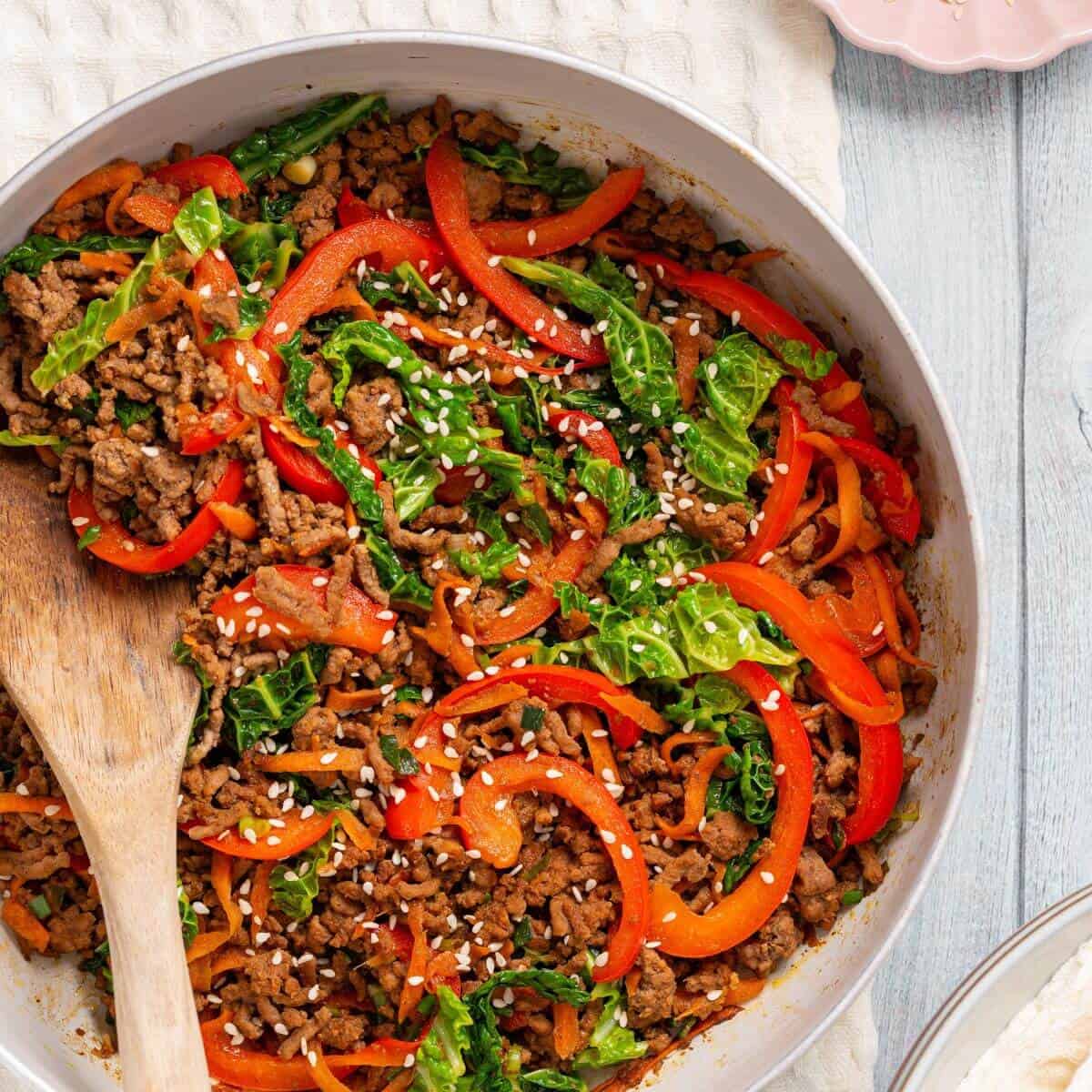 A skillet filled with a ground meat stir fry, featuring strips of red bell pepper, shredded cabbage, and sesame seeds, with a wooden spoon resting inside.