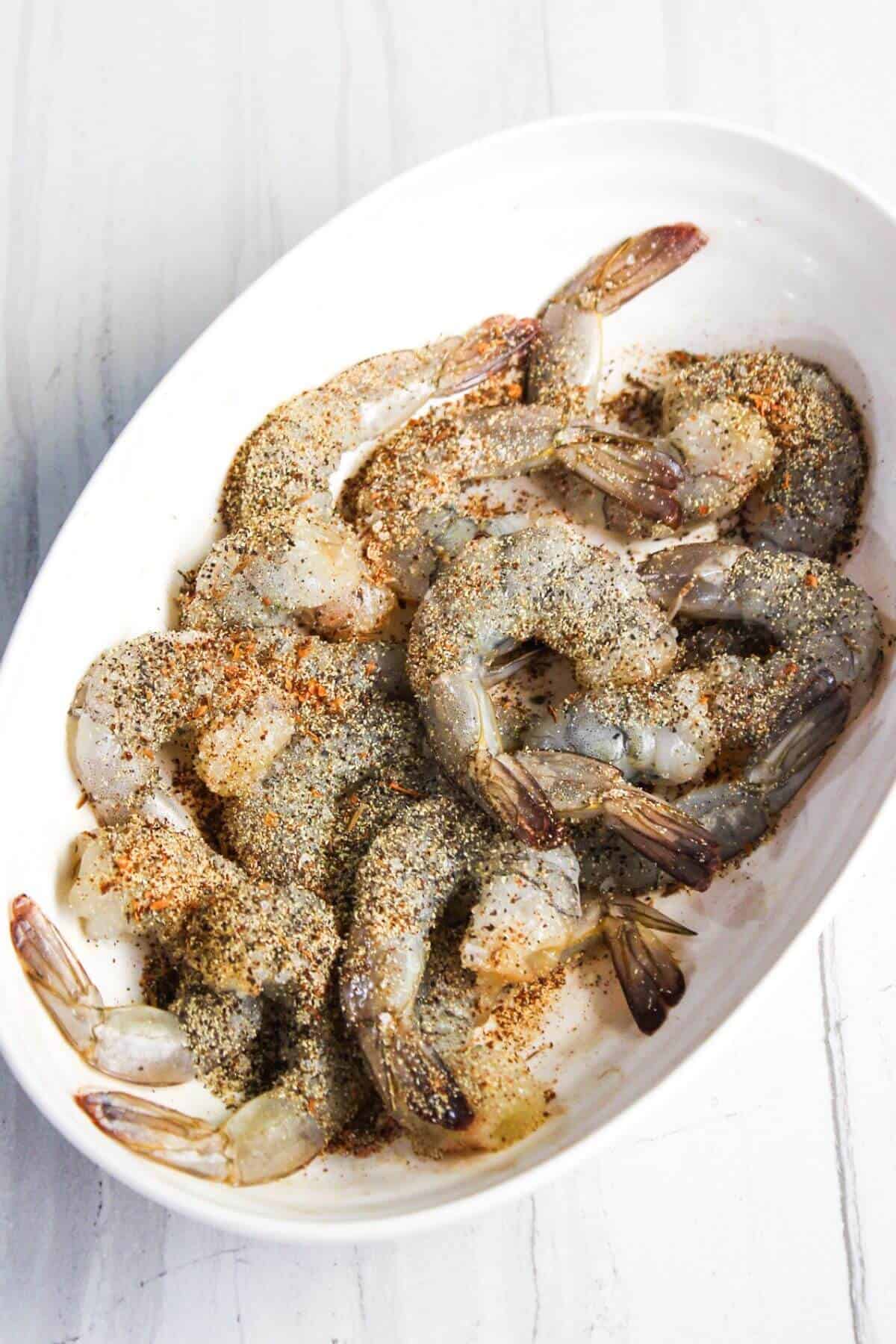 A white oval dish containing raw shrimp, seasoned with a mix of spices.