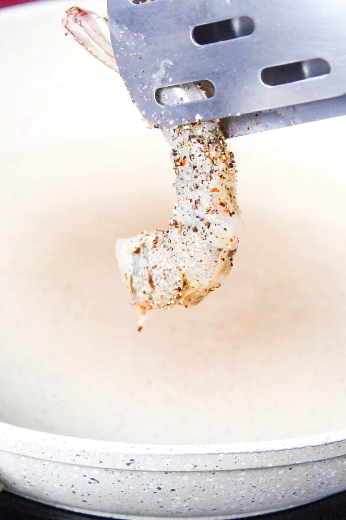 A seasoned shrimp being held by a metal spatula over a white frying pan.