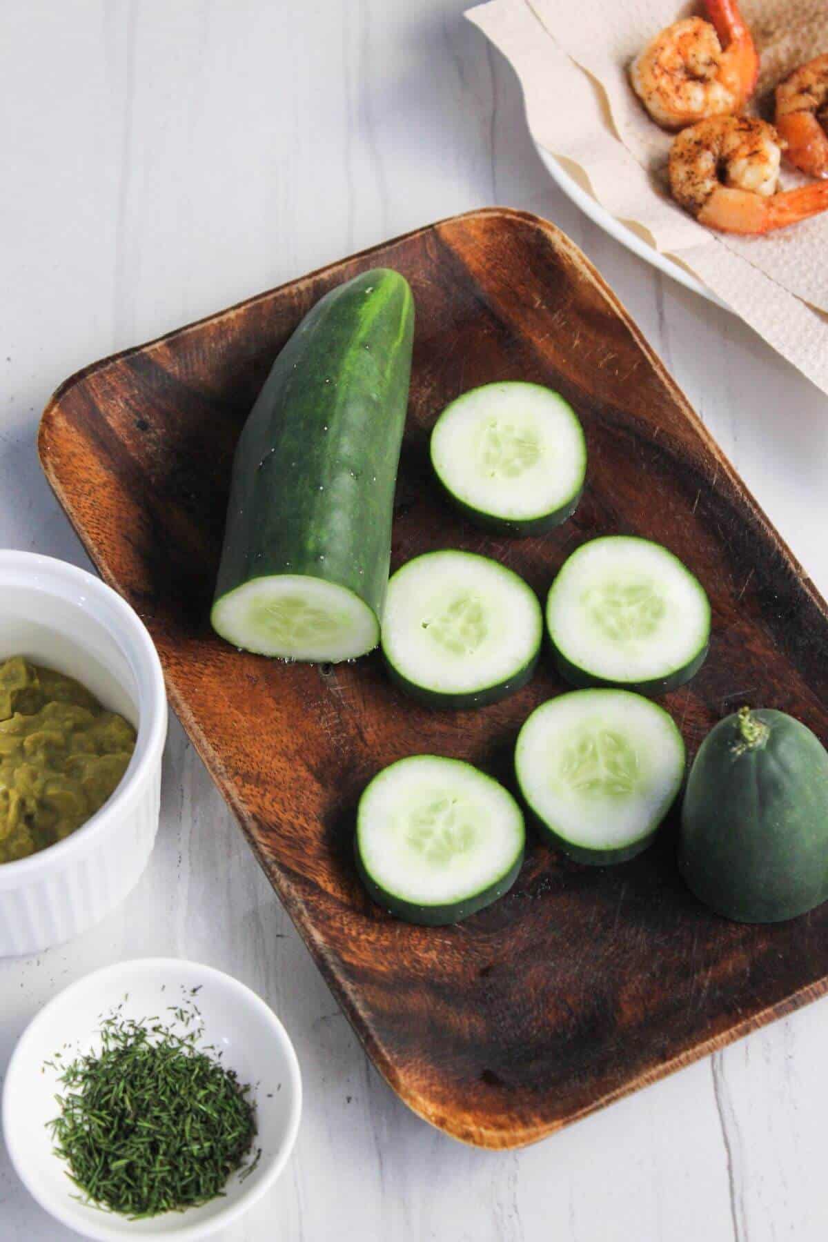 Sliced cucumber on a wooden tray, a small bowl of dill, a ramekin with green dip, and cooked shrimps on a paper towel.