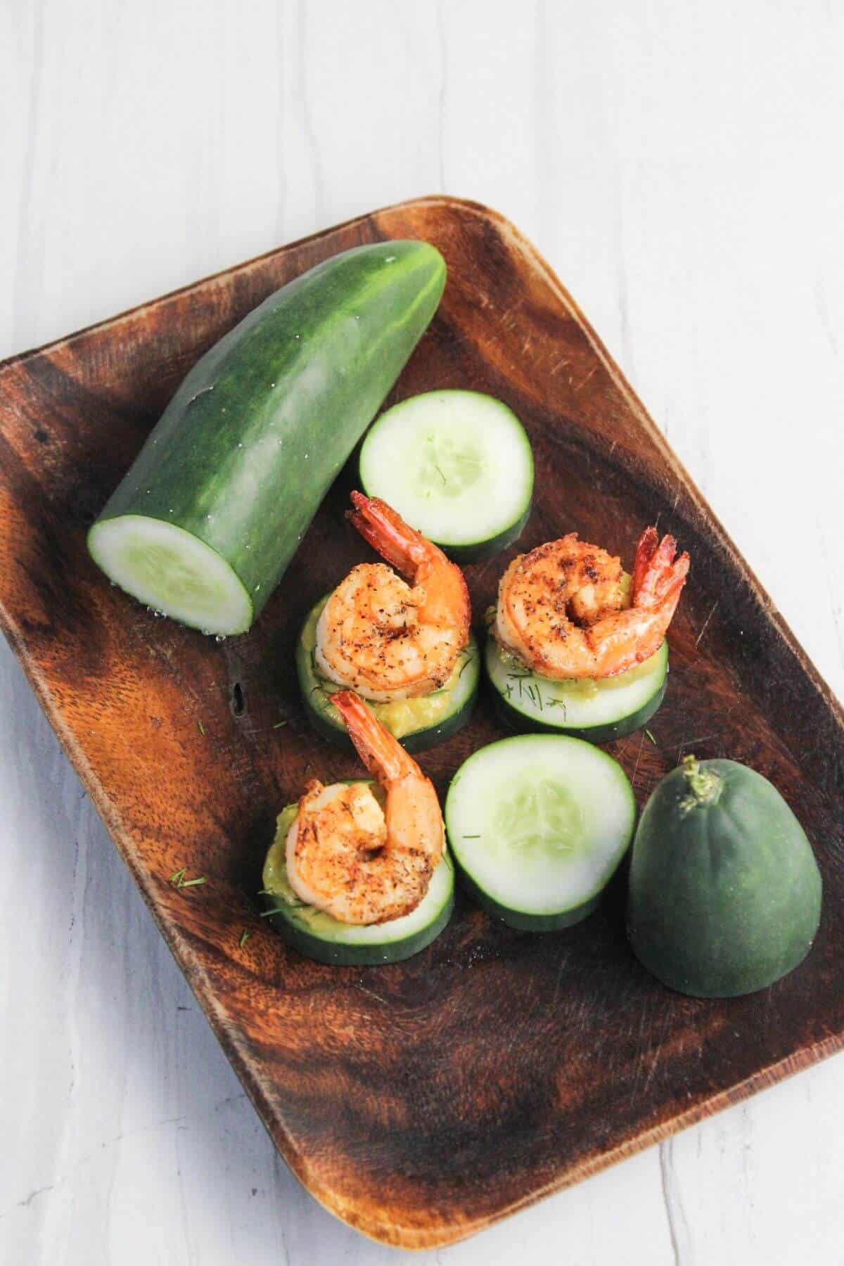 A wooden tray holds a sliced cucumber, two unsliced cucumbers, and three pieces of shrimp placed on cucumber slices.