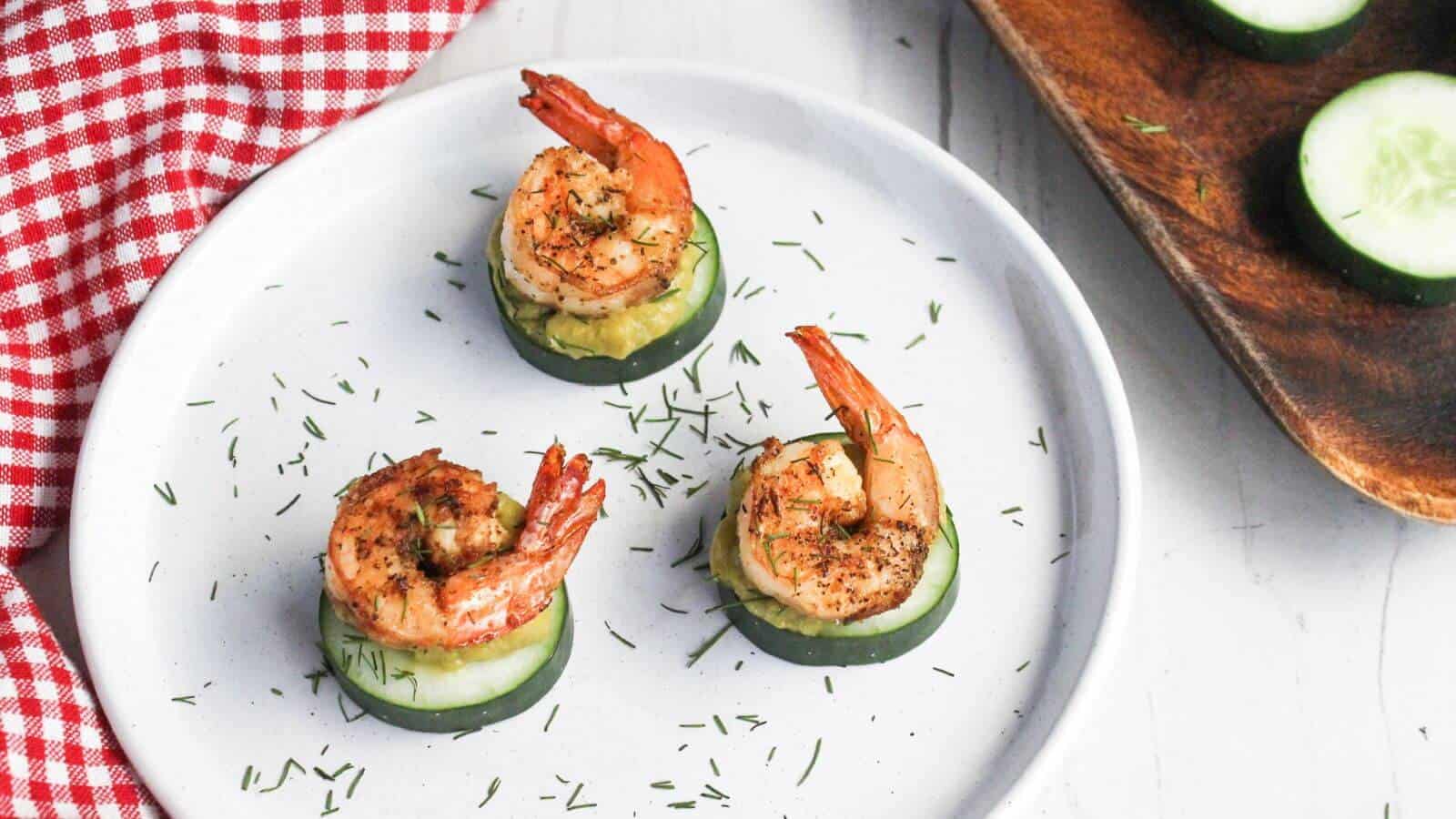 Three grilled shrimp pieces atop cucumber slices with a sprinkle of herbs, on a white plate near a red and white checkered cloth.