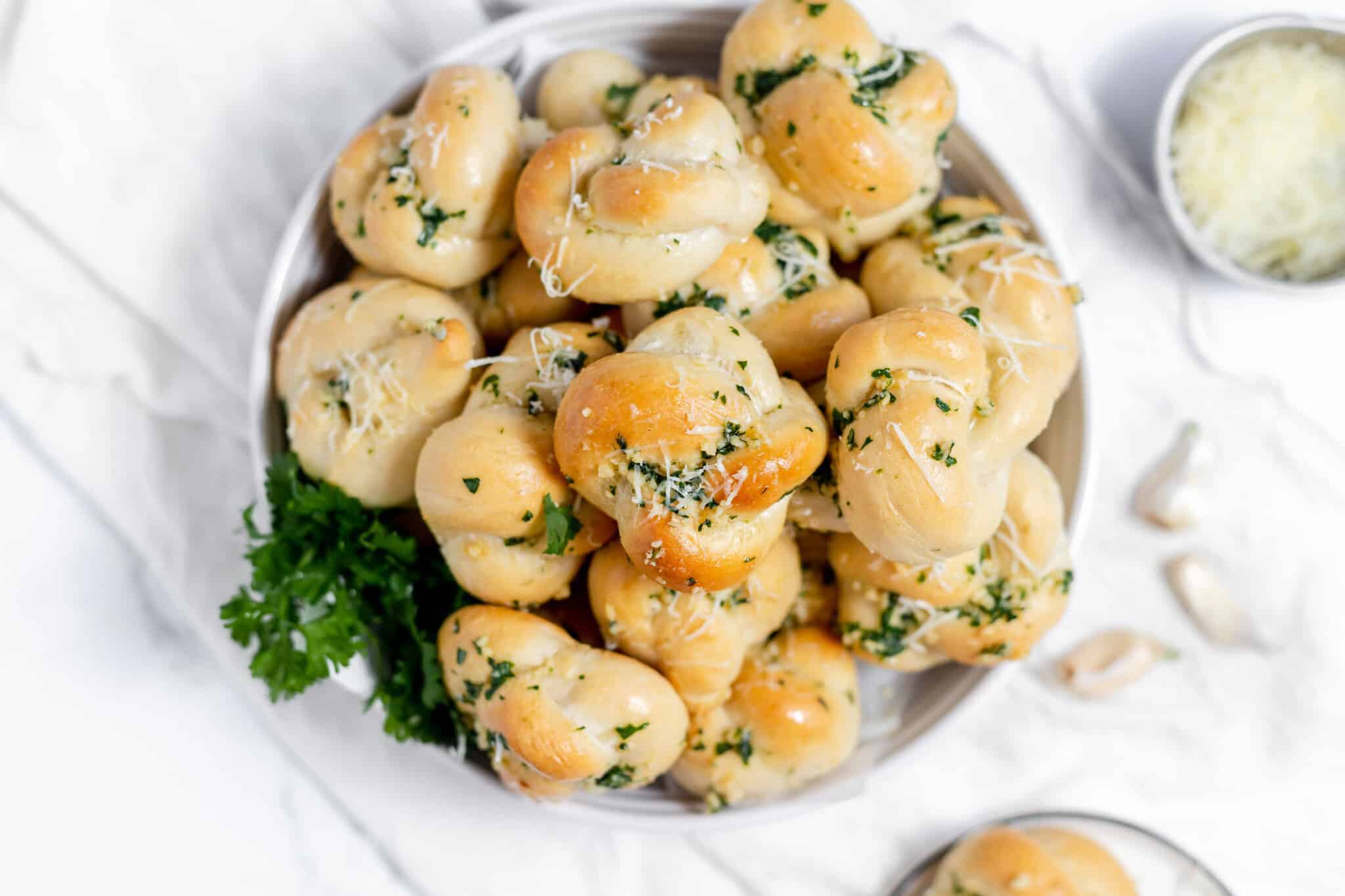 A bowl filled with garlic knots topped with herbs and grated cheese, with a garnish of fresh parsley on the side. Perfect for those easy bread recipes to satisfy every carb craving.