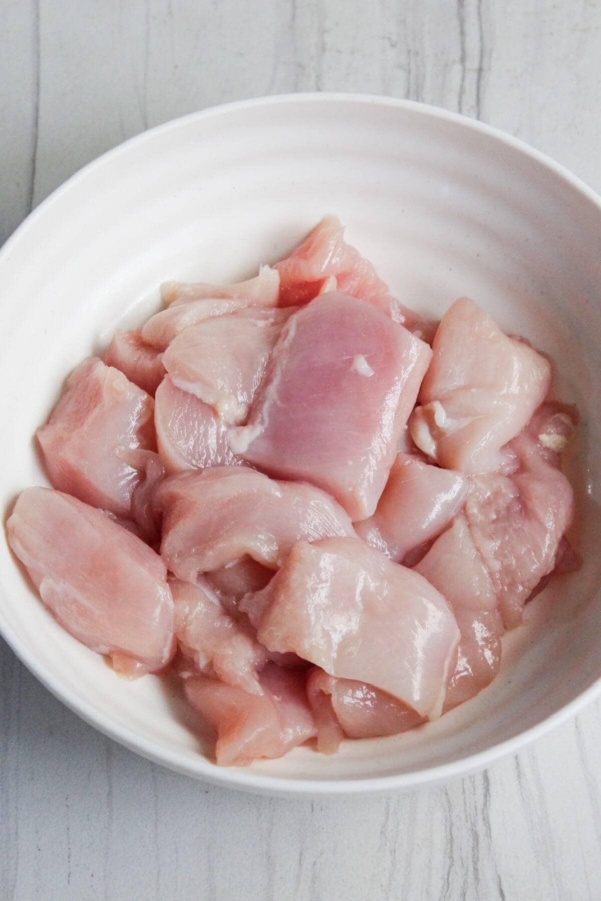 A white bowl filled with pieces of raw, pink chicken breast on a light-colored surface.