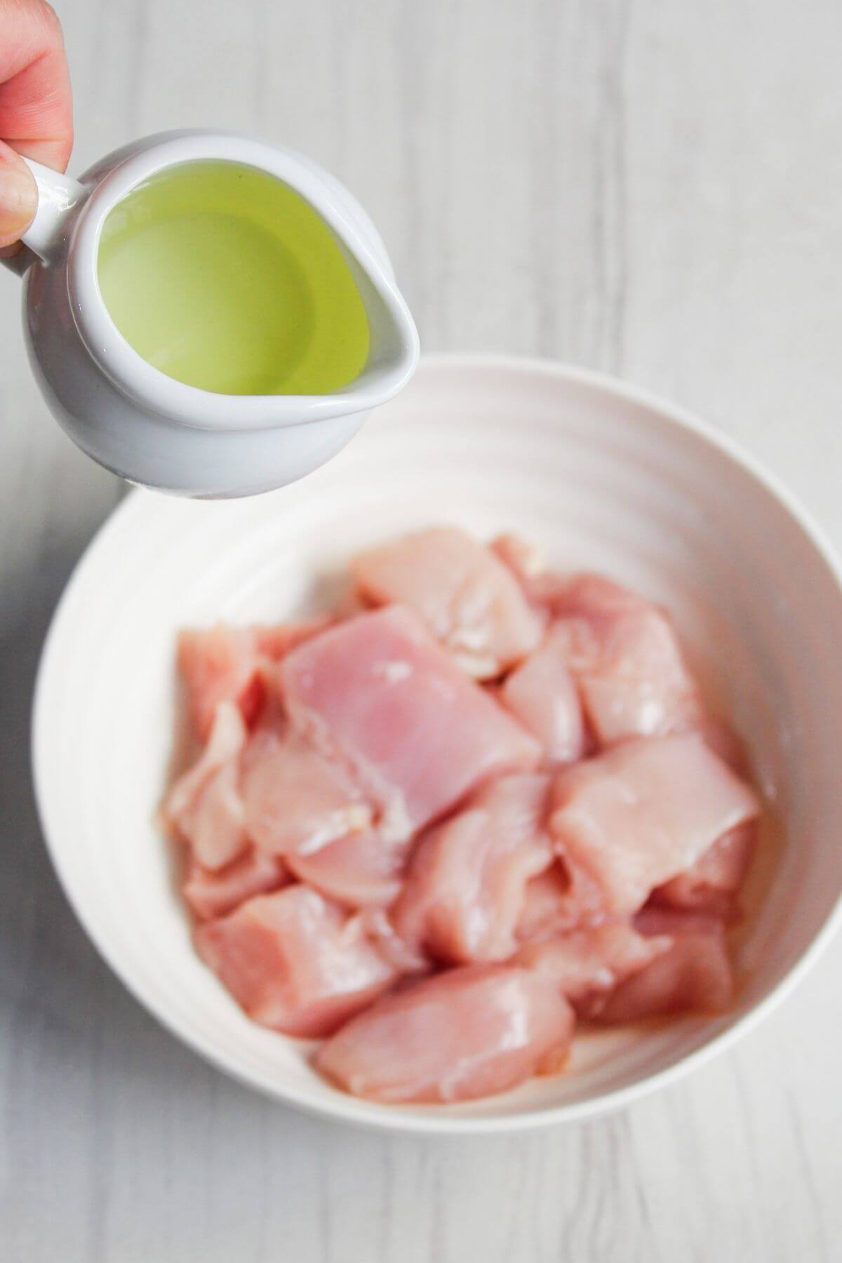 A small pitcher of oil being poured into a bowl containing raw, cubed chicken pieces.