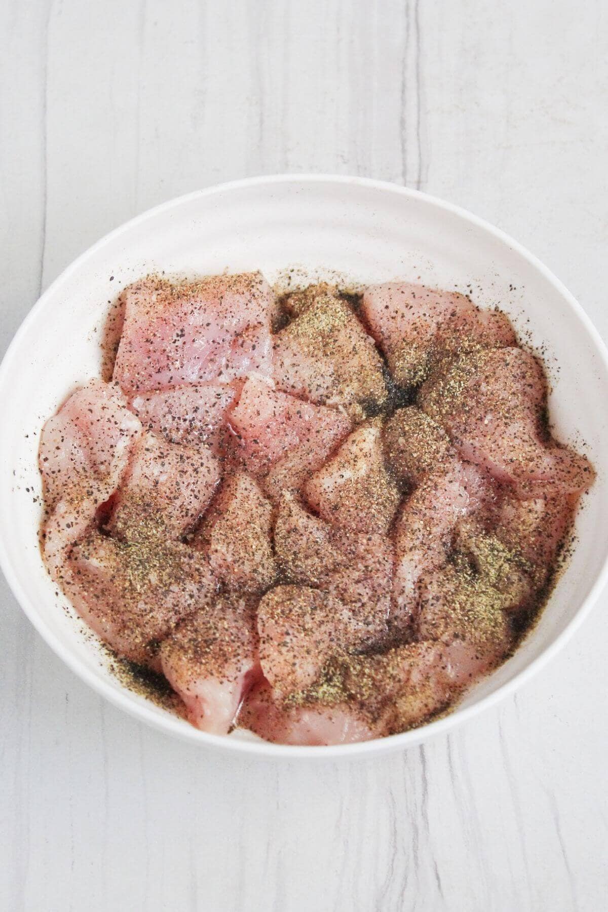 A white bowl filled with raw, cubed chicken pieces seasoned with black pepper. The bowl is placed on a light-colored surface.