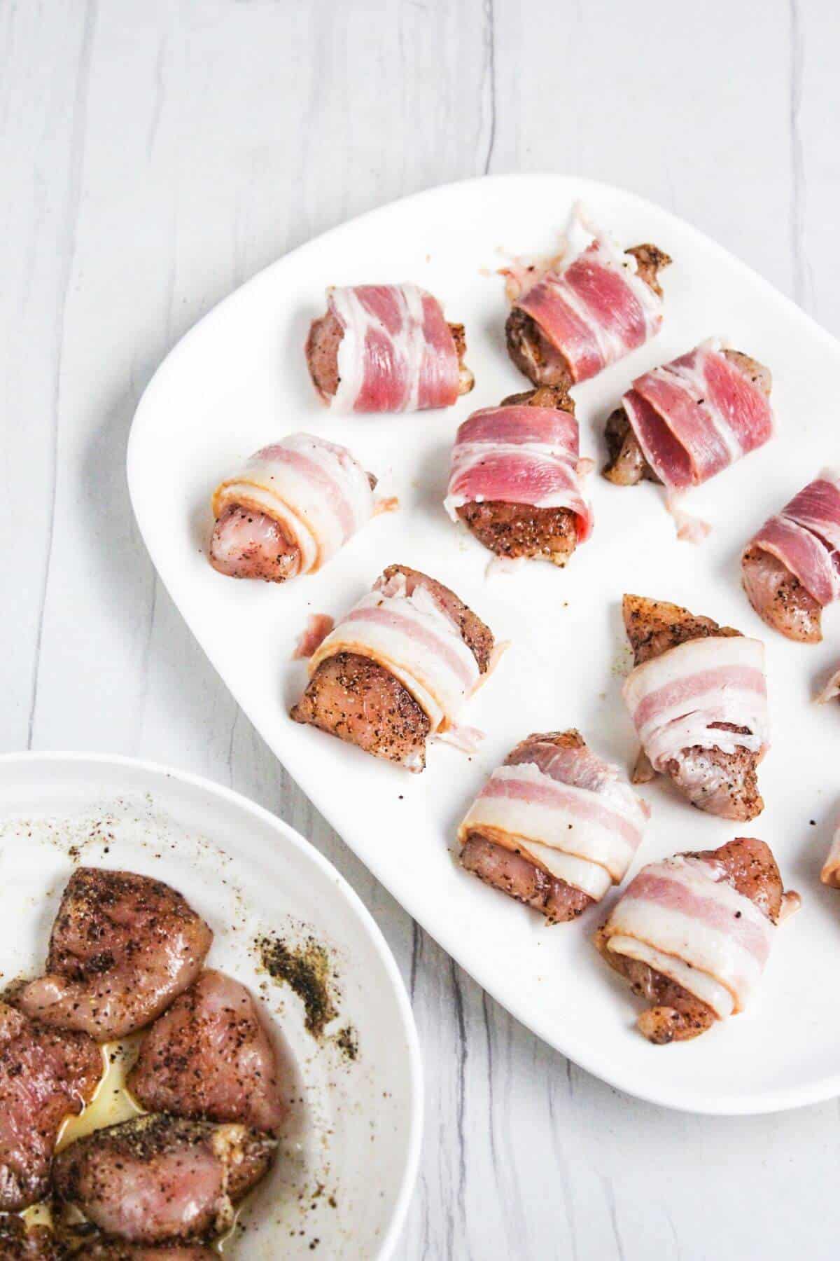 A white plate with bacon-wrapped, seasoned meat pieces on a white surface. In the bottom left, a bowl containing more seasoned meat pieces.