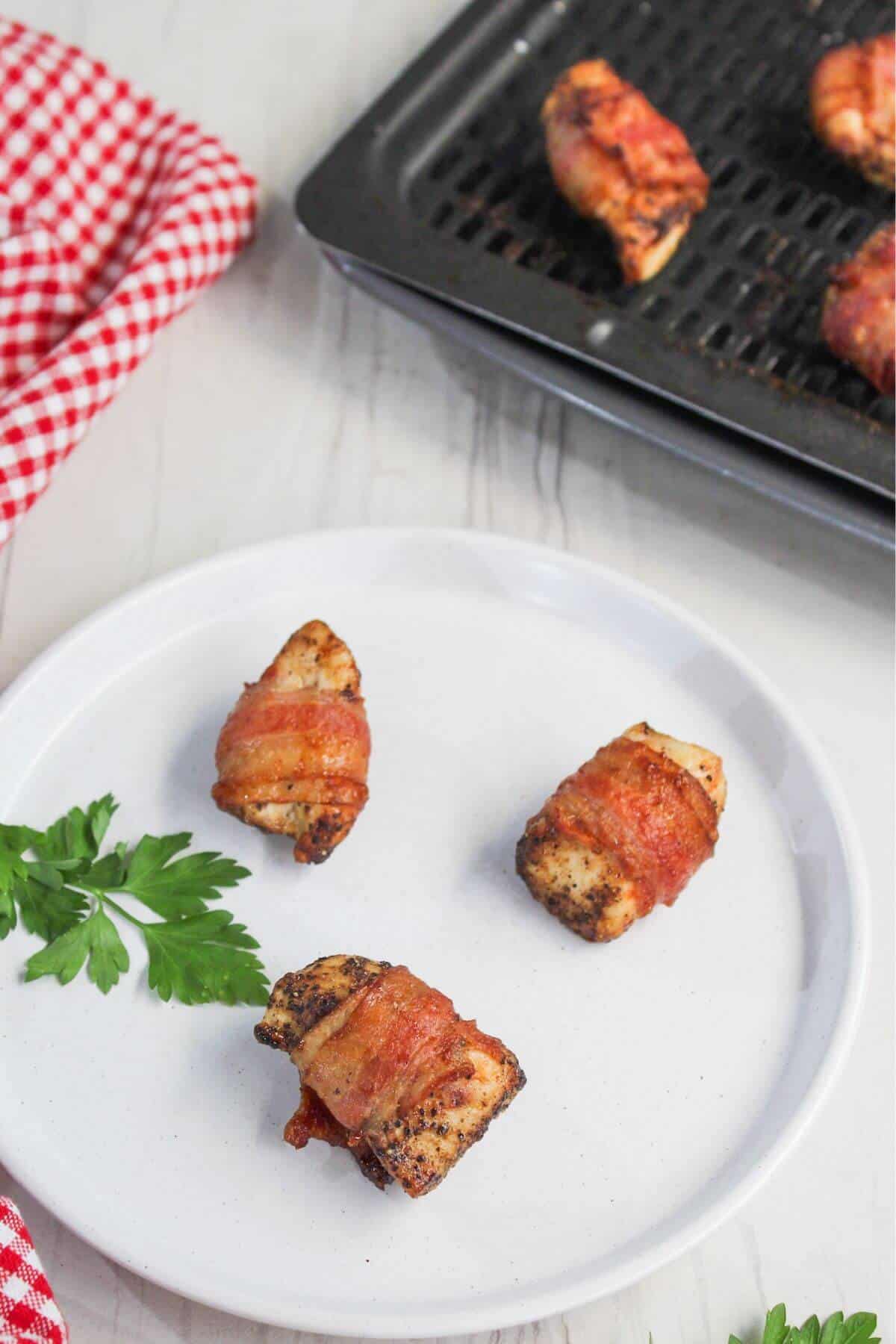 Three bacon-wrapped chicken bites on a white plate, garnished with parsley; a baking tray with more bites and a red-checkered cloth are in the background.