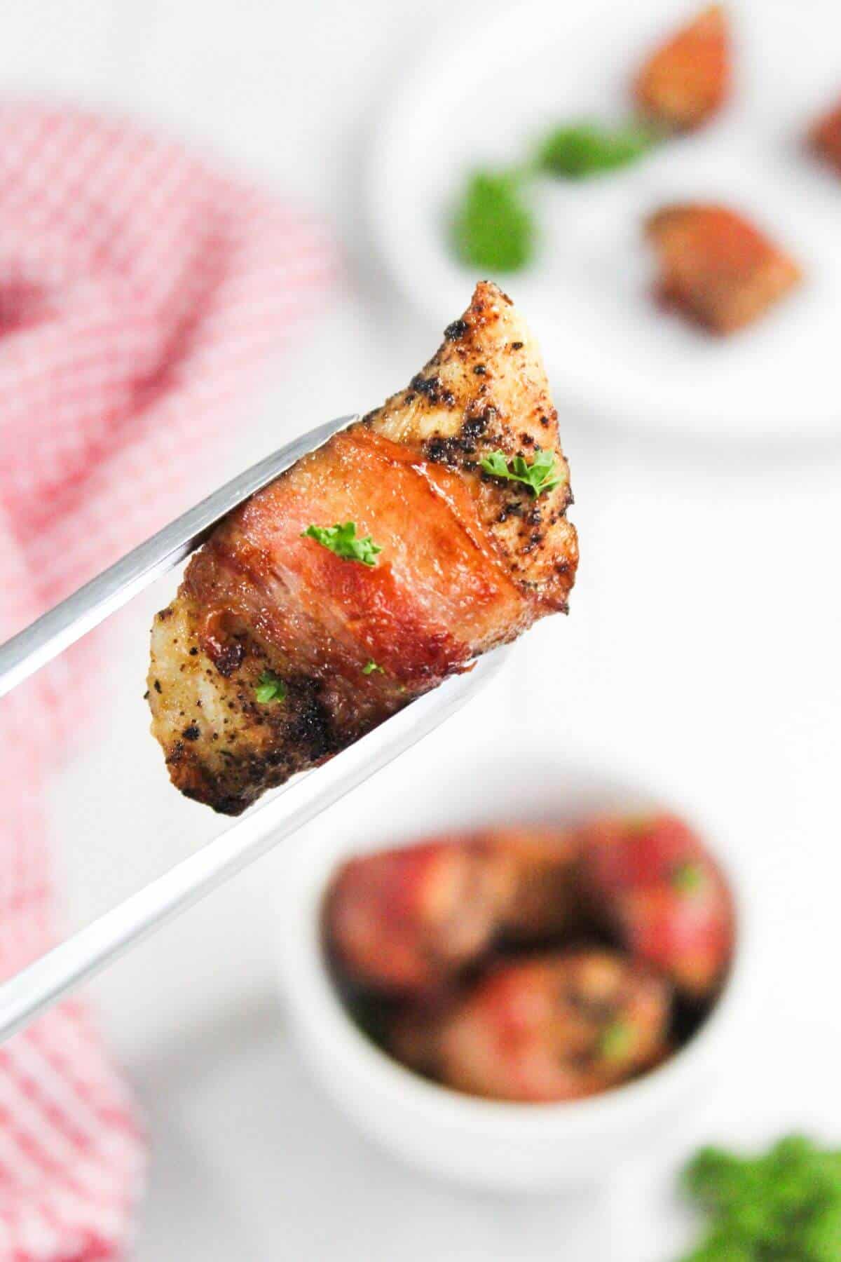 Close-up of metal tongs holding a piece of cooked meat wrapped in bacon, garnished with herbs. A blurred bowl with more pieces and a red-checked cloth are visible in the background.