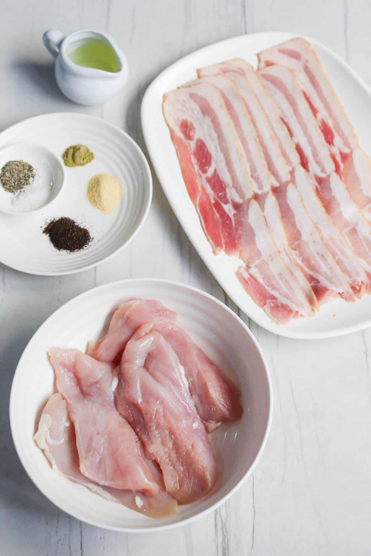 Raw chicken breasts in a white bowl, uncooked bacon strips on a plate, a small white jug of oil, and a small dish with various spices on a white surface.