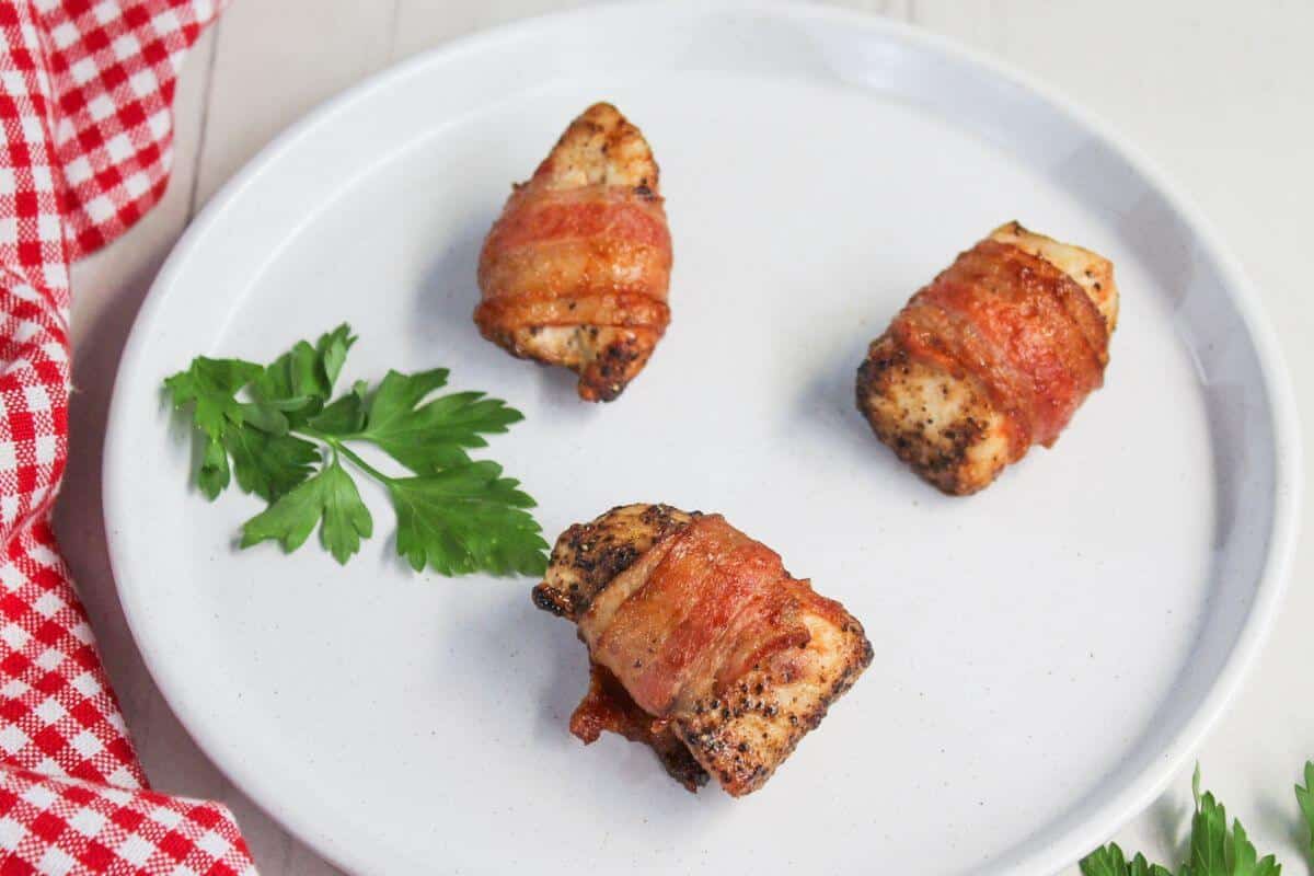 Three bacon-wrapped chicken pieces on a white plate, garnished with parsley, next to a red and white checkered cloth.