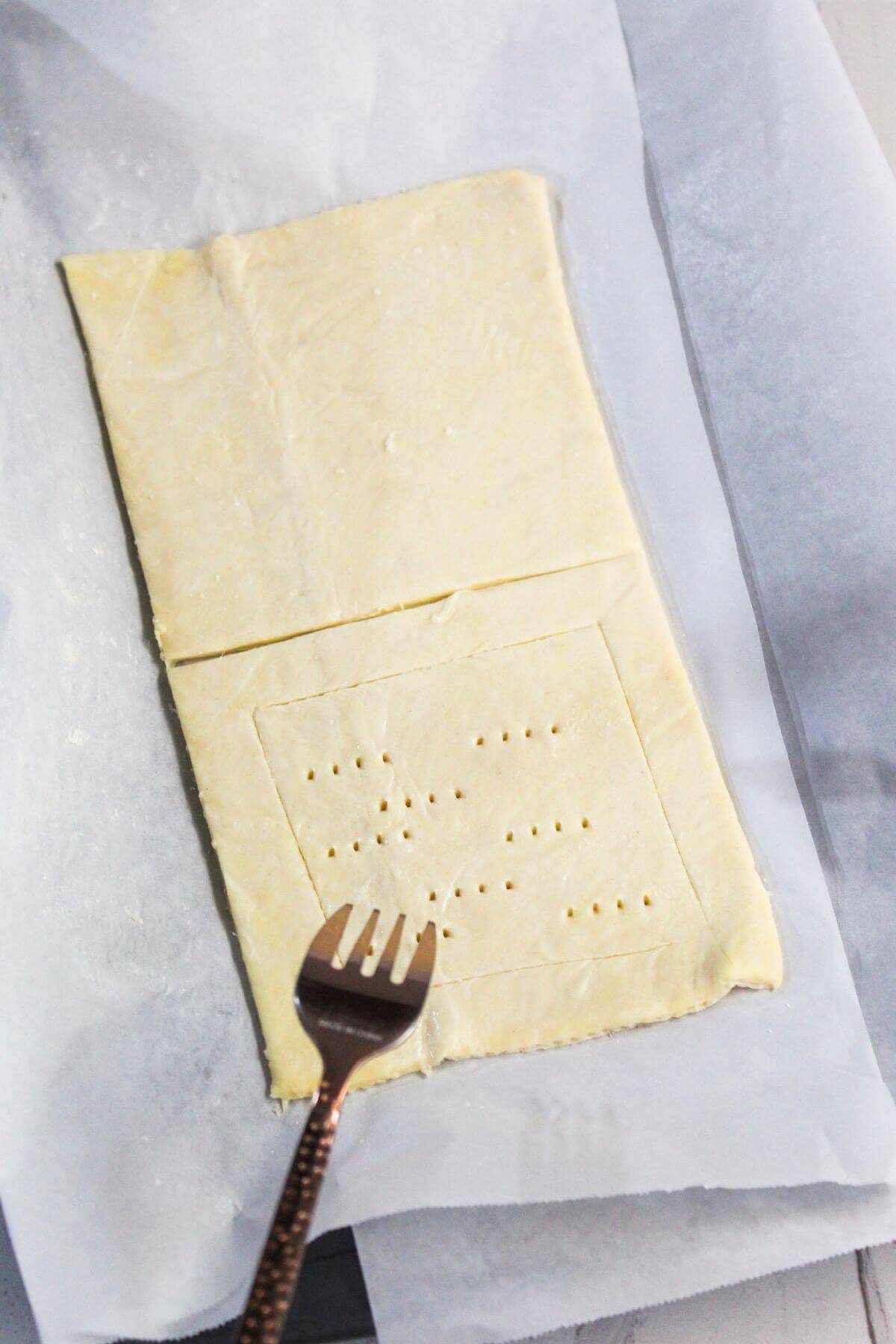 A rectangular piece of puff pastry on parchment paper, with the bottom half scored and pricked with fork holes for a cream cheese danish. A fork with a copper handle rests on the pastry.