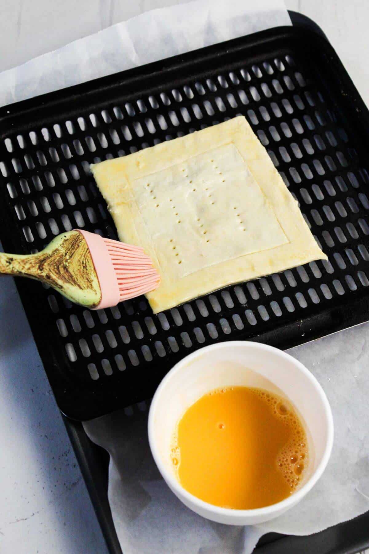 A cream cheese danish square on a black perforated baking tray is being brushed with egg wash, with a white bowl containing egg wash next to it.