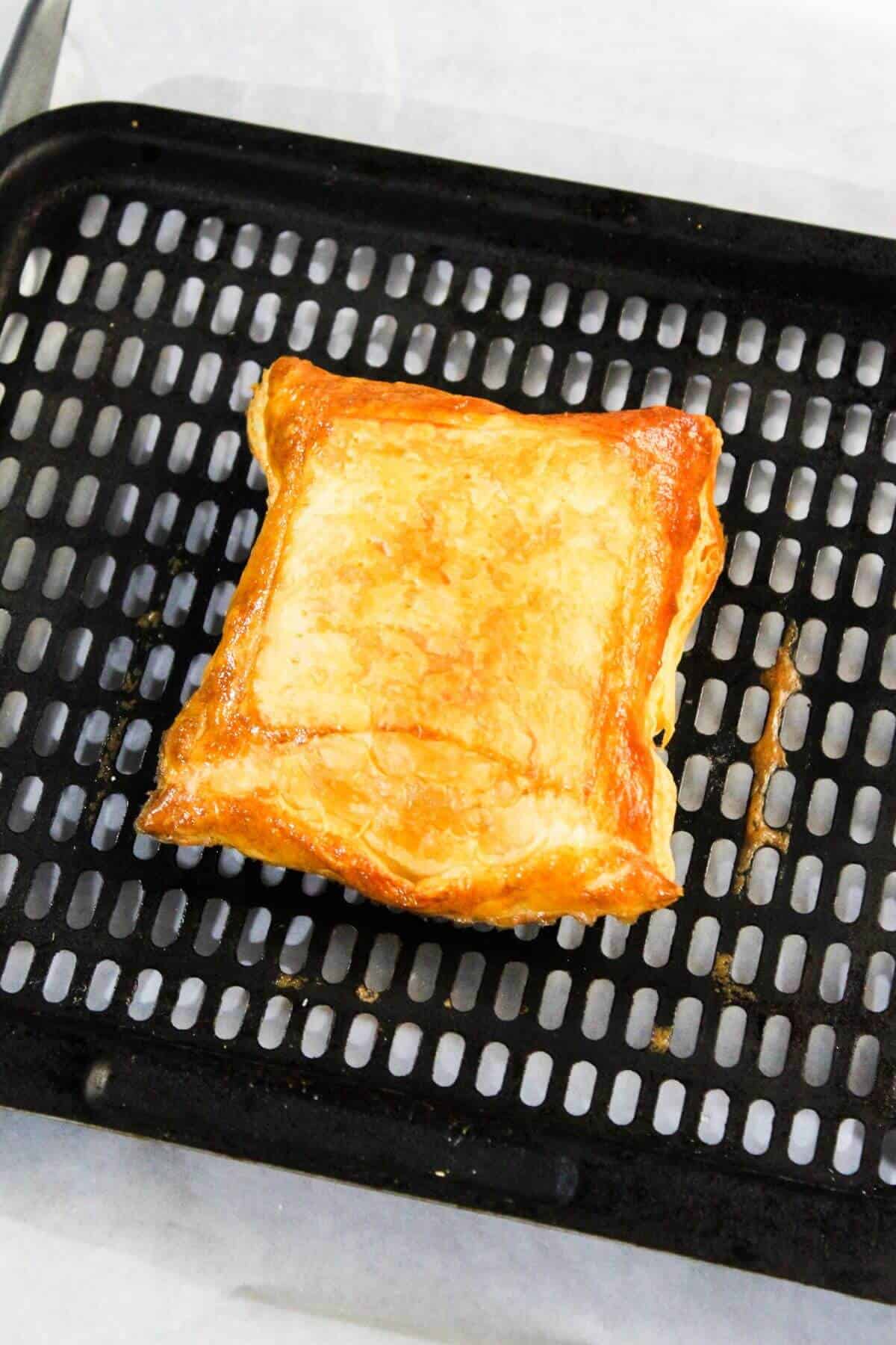 A golden-brown, flaky cream cheese danish rests on a black perforated baking sheet.