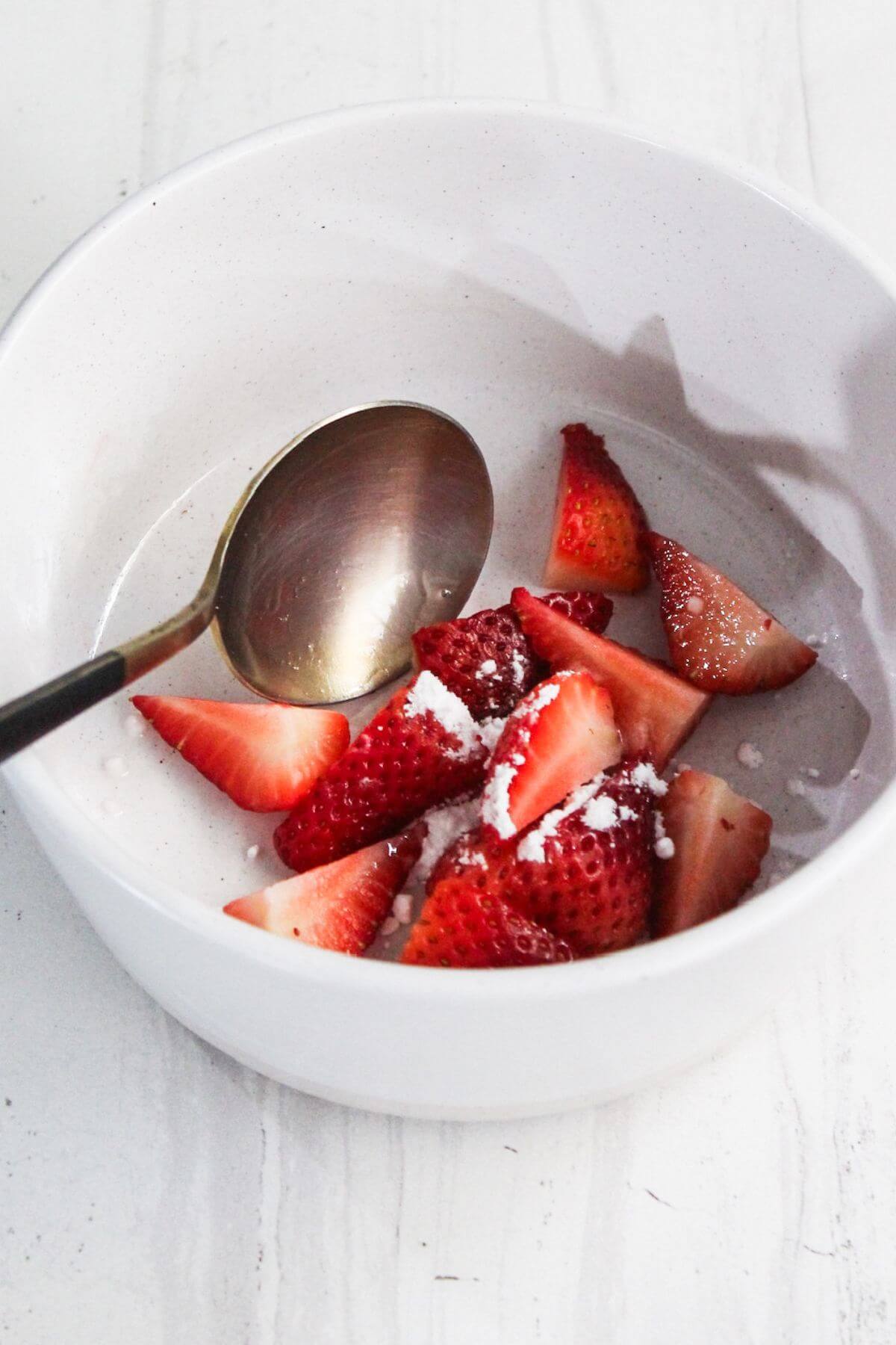 A white bowl containing cut strawberries sprinkled with sugar sits invitingly. A metal spoon is partially submerged among the strawberries, evoking the richness of a cream cheese Danish.