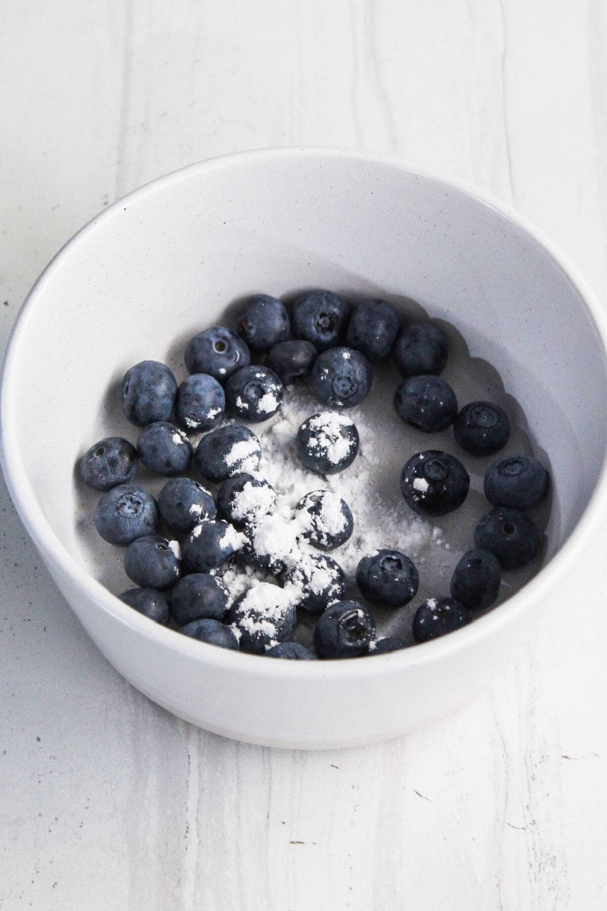 A white bowl containing fresh blueberries, some lightly dusted with powdered sugar, sits on a light-colored surface, reminiscent of the toppings on a delectable cream cheese danish.