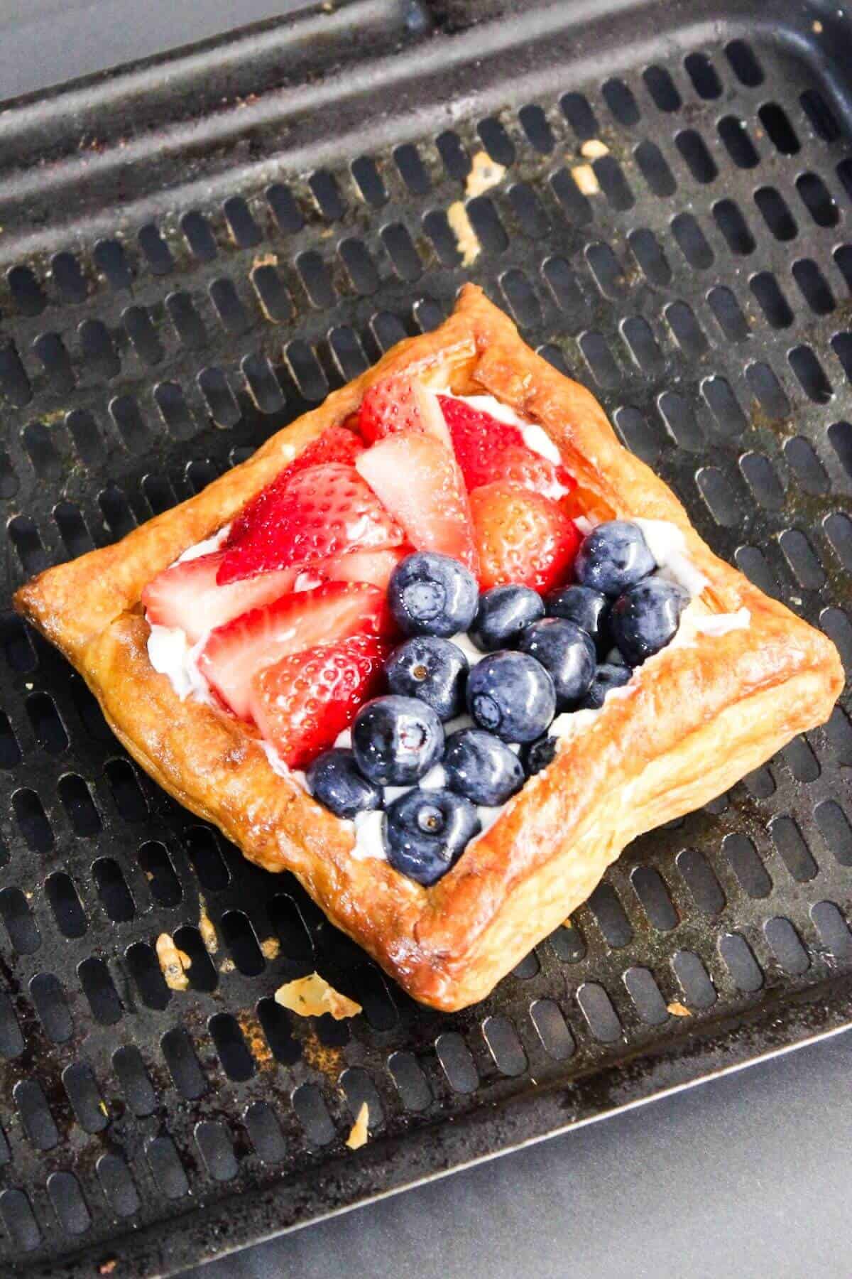A square puff pastry topped with sliced strawberries and blueberries resembling a classic cream cheese danish rests on a dark, perforated baking tray.
