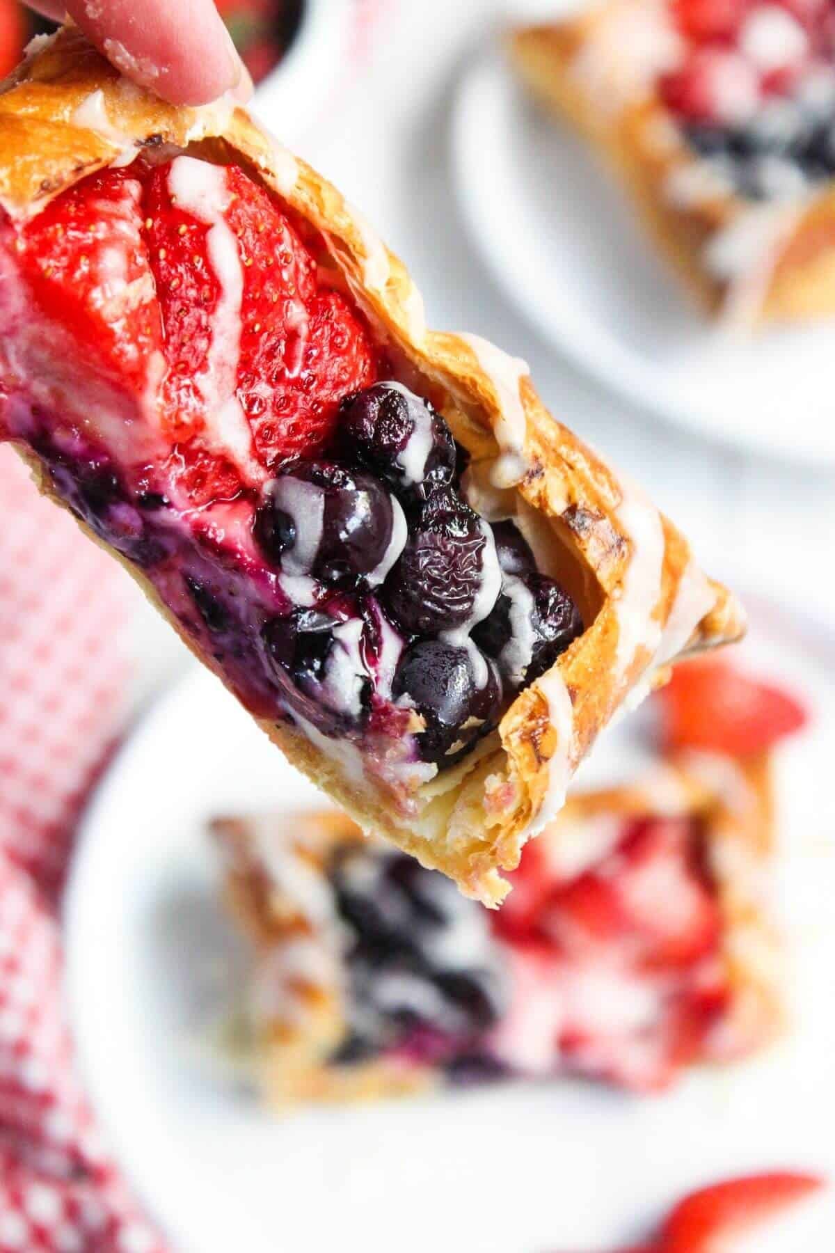 A close-up of a hand holding a cream cheese danish topped with strawberries, blueberries, and icing.