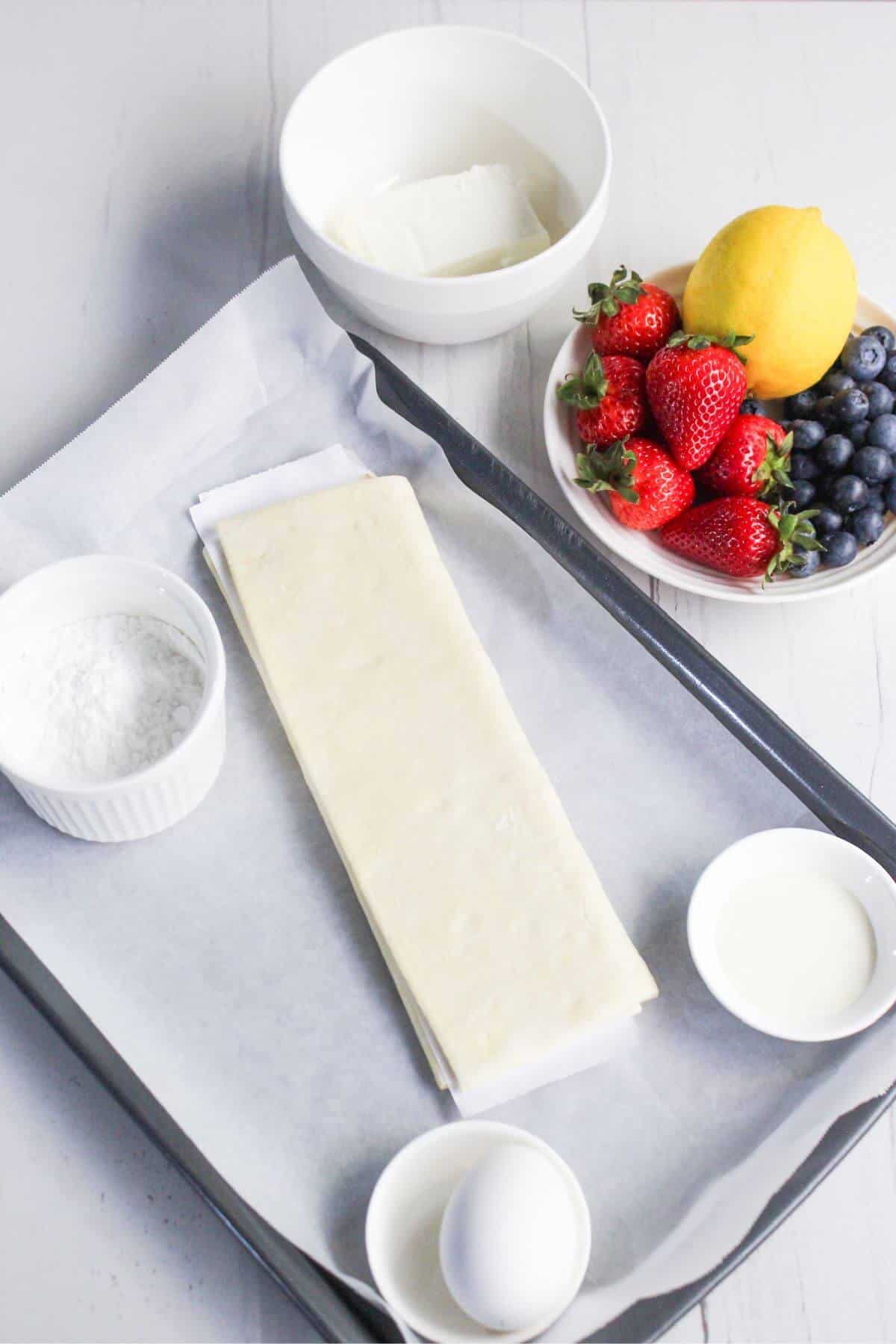 A baking sheet with parchment paper holds a rectangle of dough, surrounded by ingredients for a cream cheese danish: a bowl of cream cheese, a lemon, strawberries, blueberries, powdered sugar, an egg, and a small bowl of cream.
