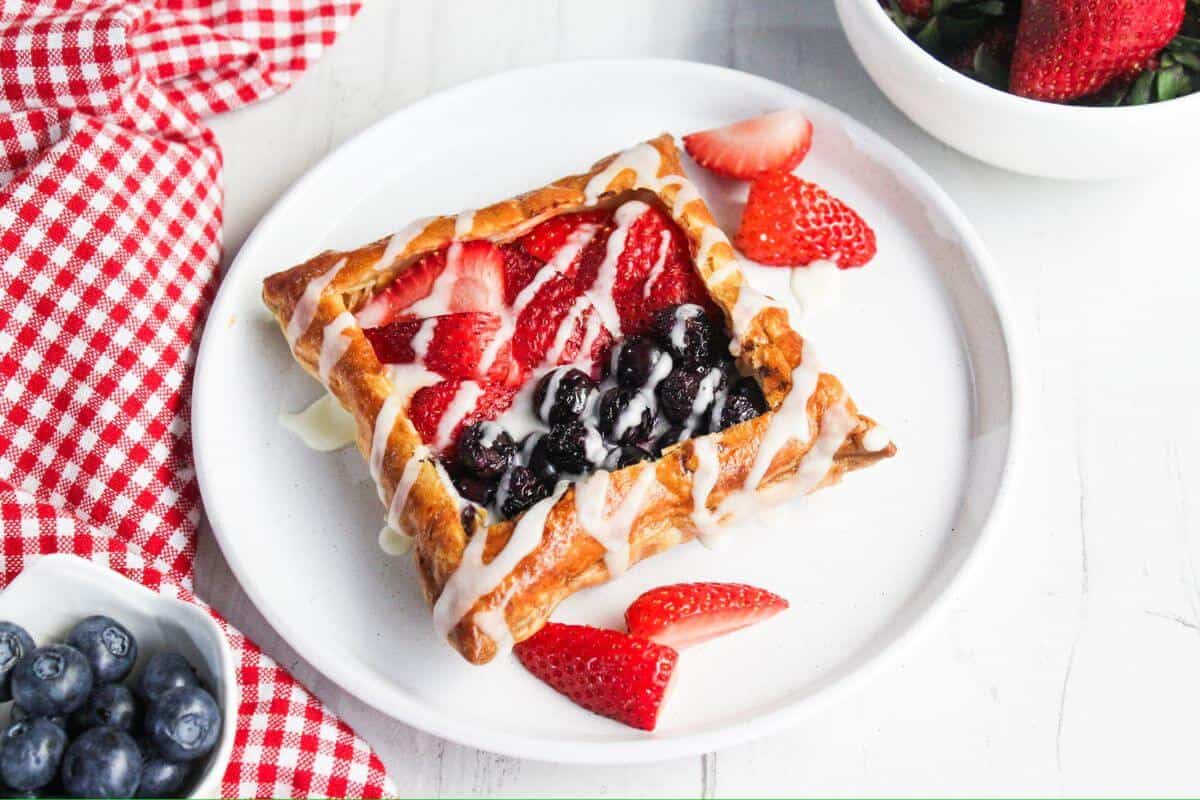 A square cream cheese danish topped with sliced strawberries and blueberries, drizzled with icing, is on a white plate. A red-checkered cloth and bowls of blueberries and strawberries are in the background.