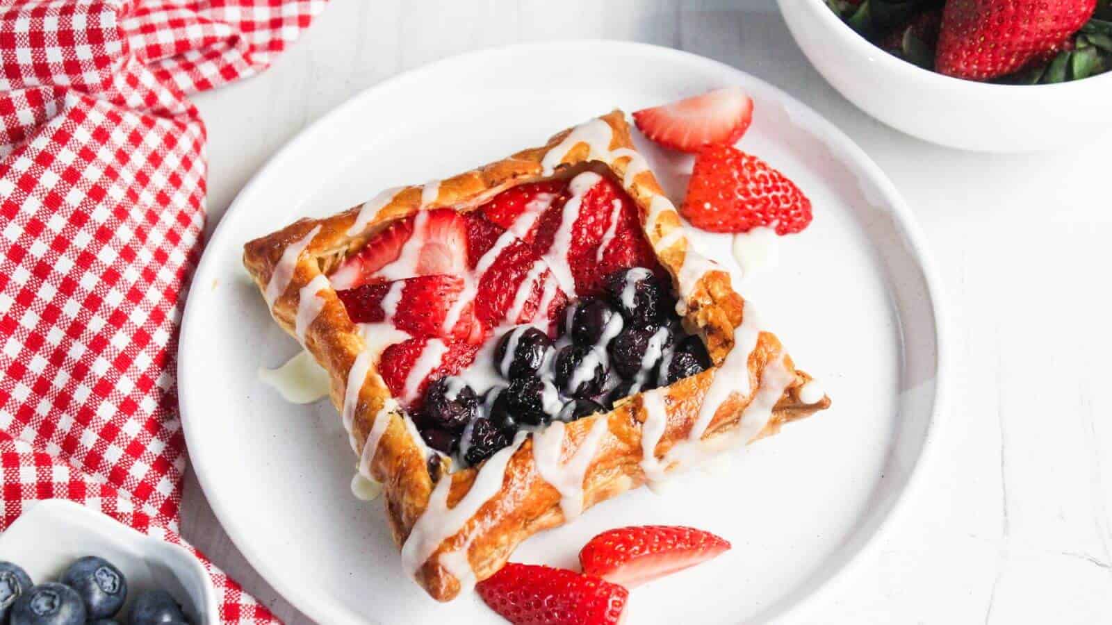 A cream cheese danish topped with strawberries and blueberries drizzled with icing is on a white plate. A red checkered cloth, a bowl of strawberries, and a small bowl of blueberries are nearby.