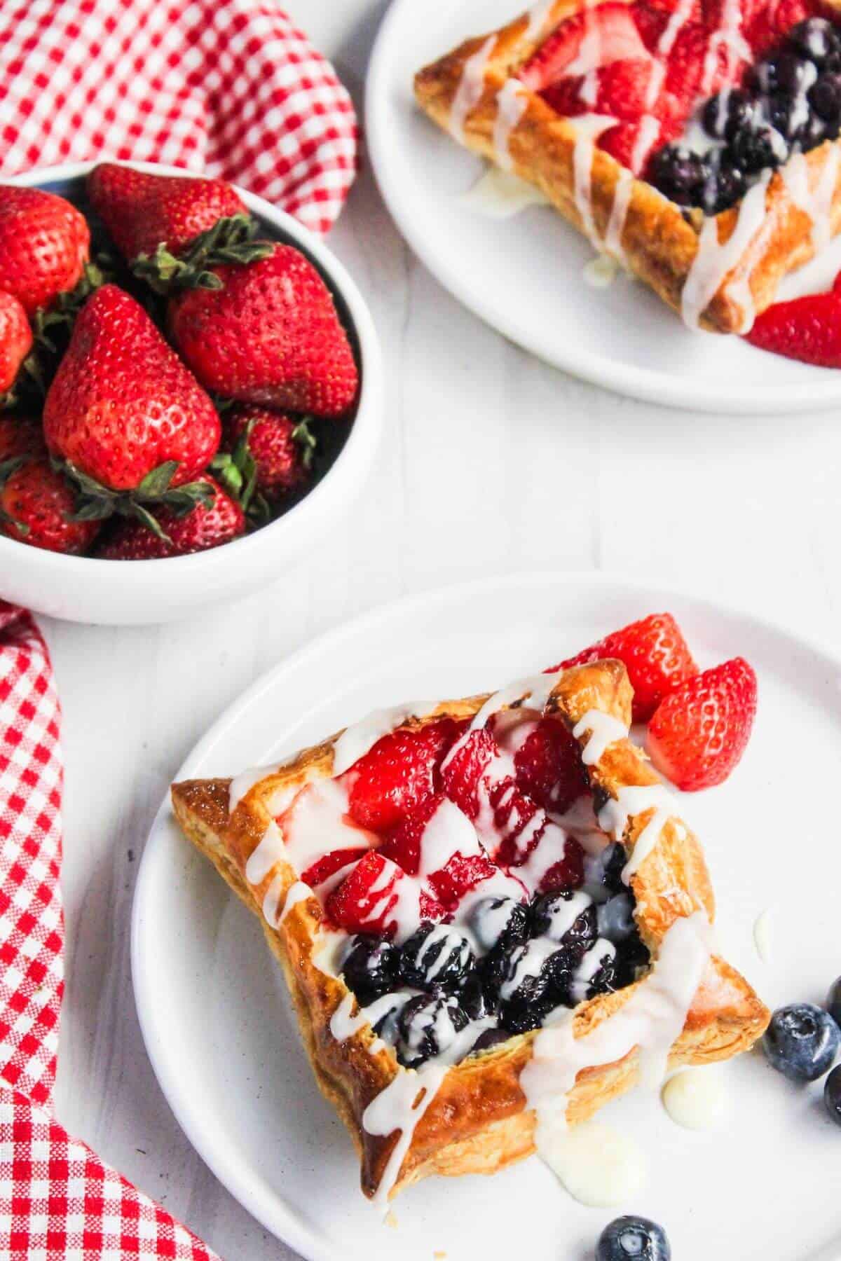 Two fruit pastries with strawberries and blueberries drizzled with icing on white plates; a bowl of strawberries and a red checkered cloth are nearby, evoking the inviting aroma of a cream cheese danish.