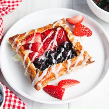 A cream cheese danish square topped with sliced strawberries and blueberries, drizzled with white icing, sits on a white plate. Sliced strawberries frame the pastry.