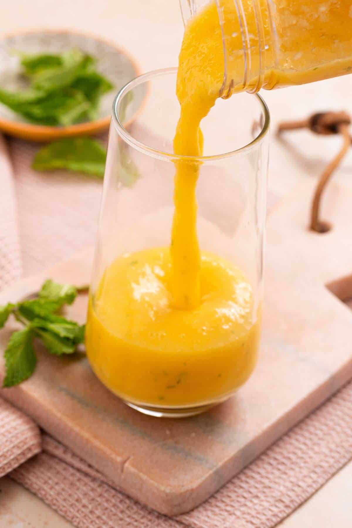 A glass is being filled with a thick, yellow mango smoothie from a jug. Mint leaves and a pink cloth are in the background.