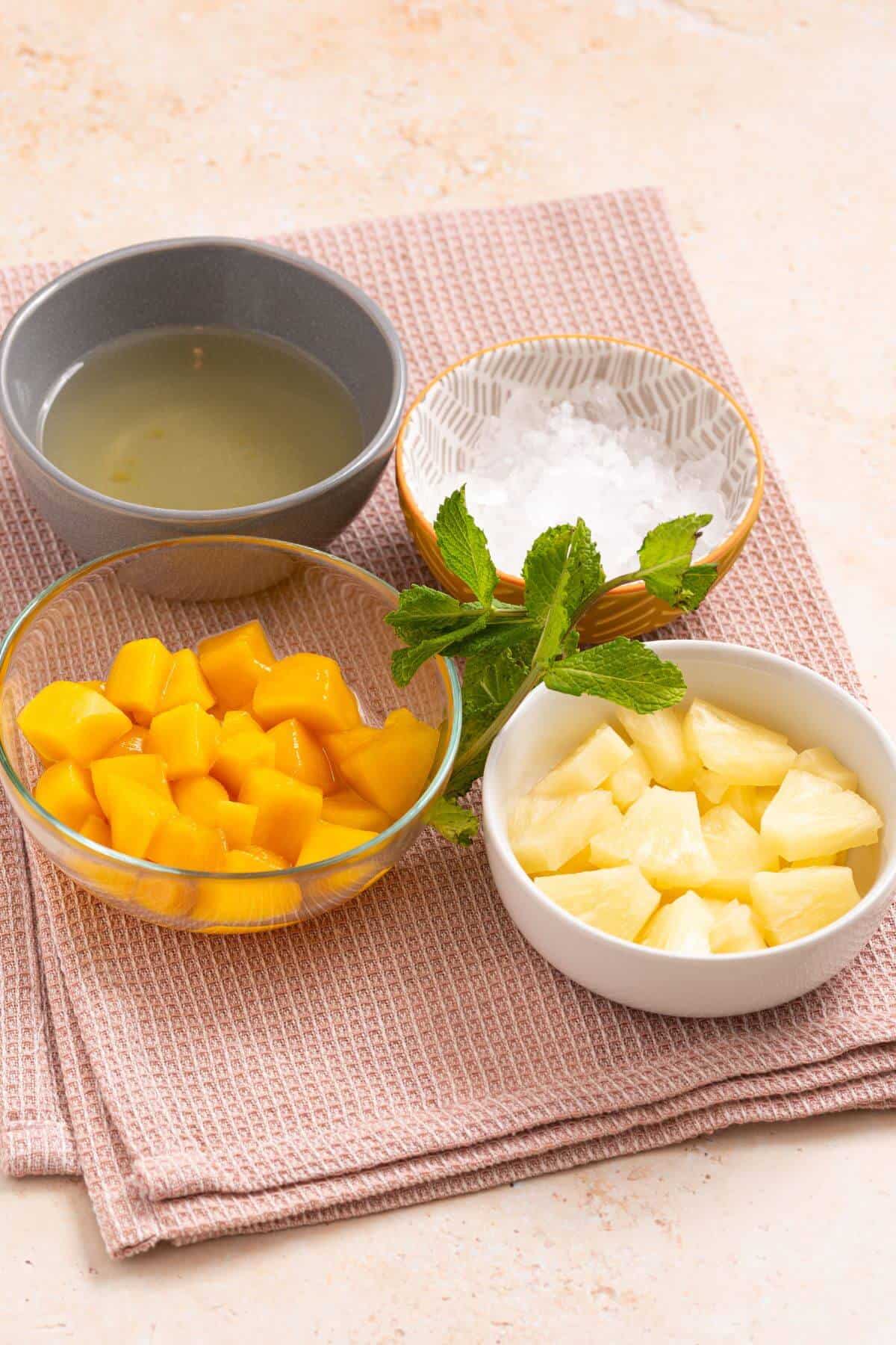 Four bowls containing lime juice, diced mango, pineapple chunks, and salt, along with a sprig of mint, are arranged on a pink textured cloth.