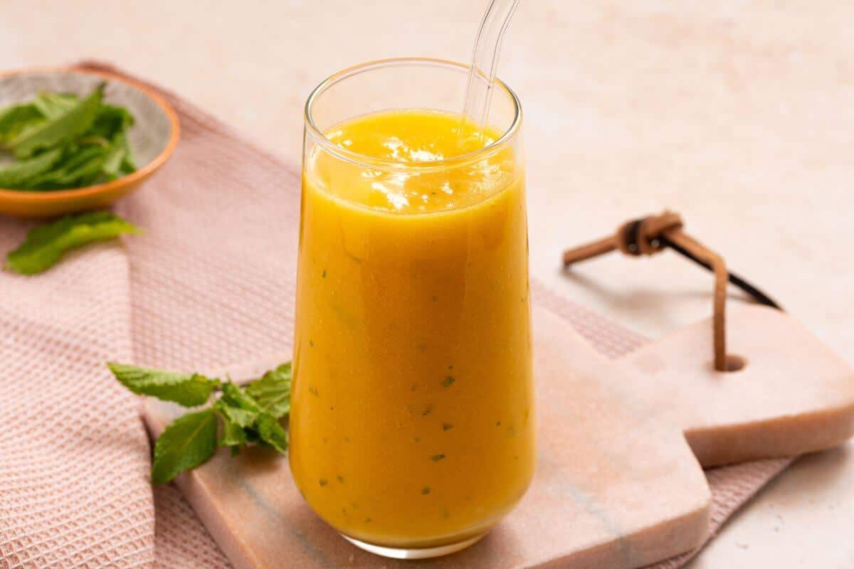 A glass of mango smoothie with a glass straw sits on a cutting board. Fresh mint leaves and a small bowl are in the background.