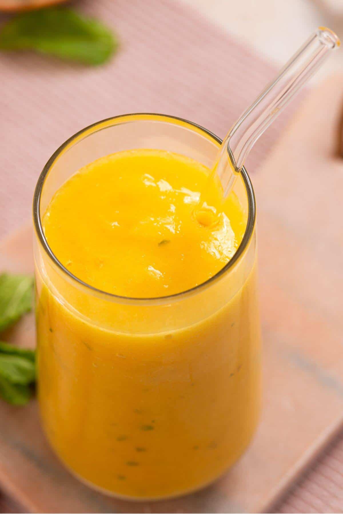 A clear glass filled with a thick, bright yellow smoothie and a clear straw, placed on a pastel pink surface with a green leaf in the background.