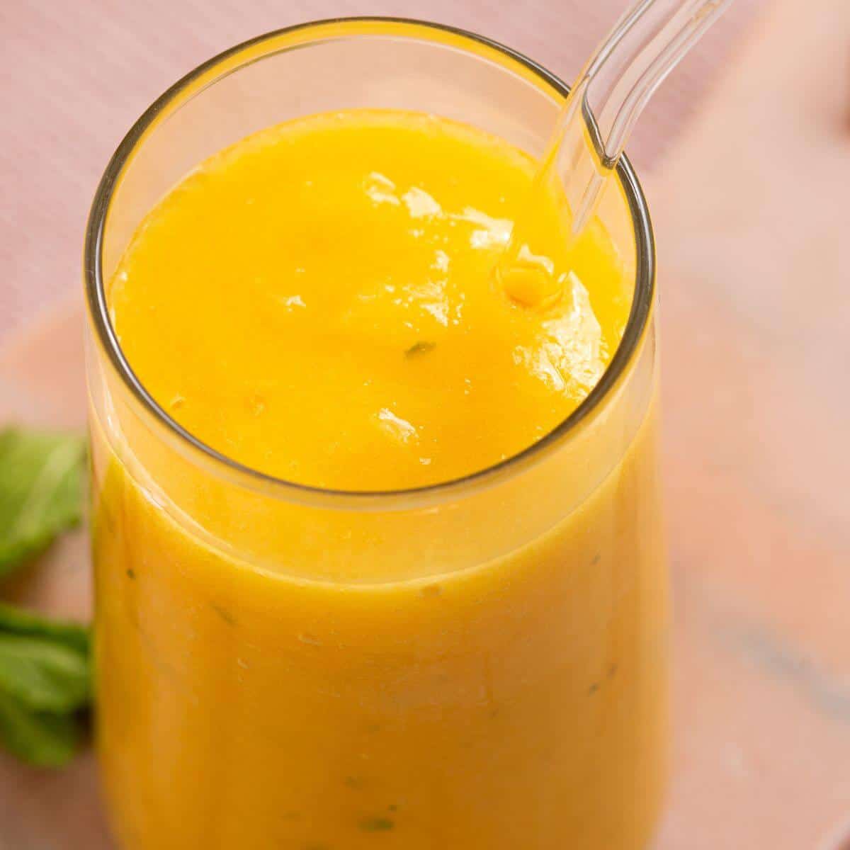 A glass filled with vibrant yellow mango lassi, containing small green herb flecks, with a clear straw inserted. A basil leaf is partially visible behind the glass.