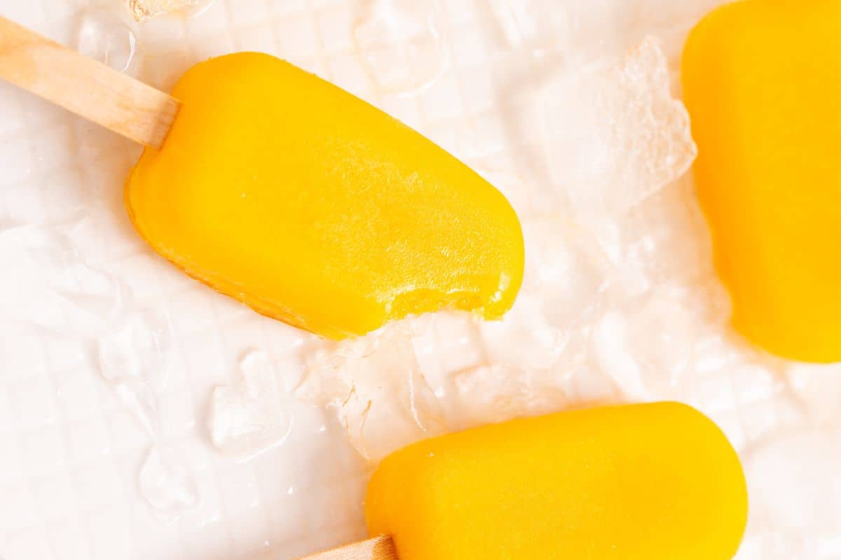 Four mango popsicles with wooden sticks, one with a bite taken out of it, placed on a light-colored surface with ice cubes scattered around.