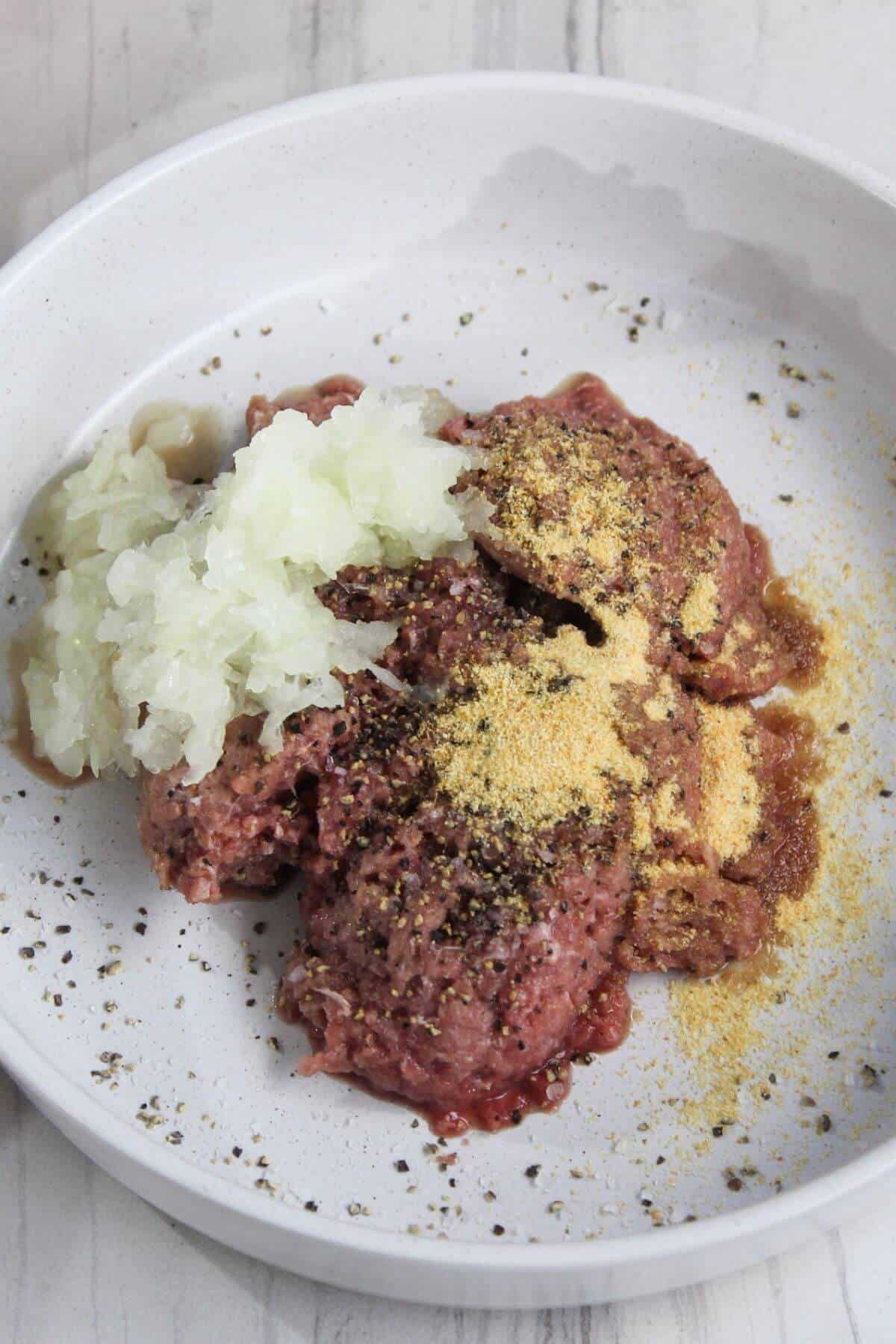 A white plate with raw ground meat, chopped onions, pepper, and seasoning powder.