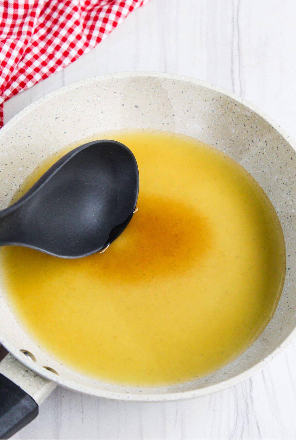 A black ladle dipping into a light yellow liquid in a white frying pan with a red and white checkered cloth in the background.