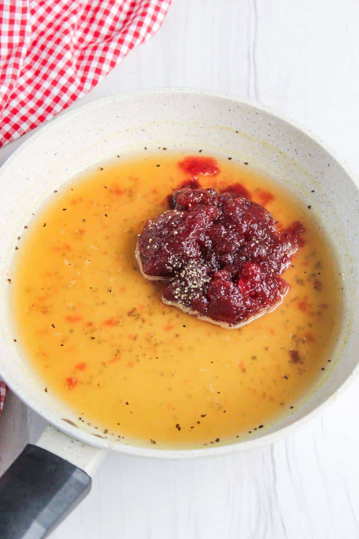 A saucepan with a yellowish liquid and a dollop of red sauce, placed on a white surface near a red and white checkered cloth.