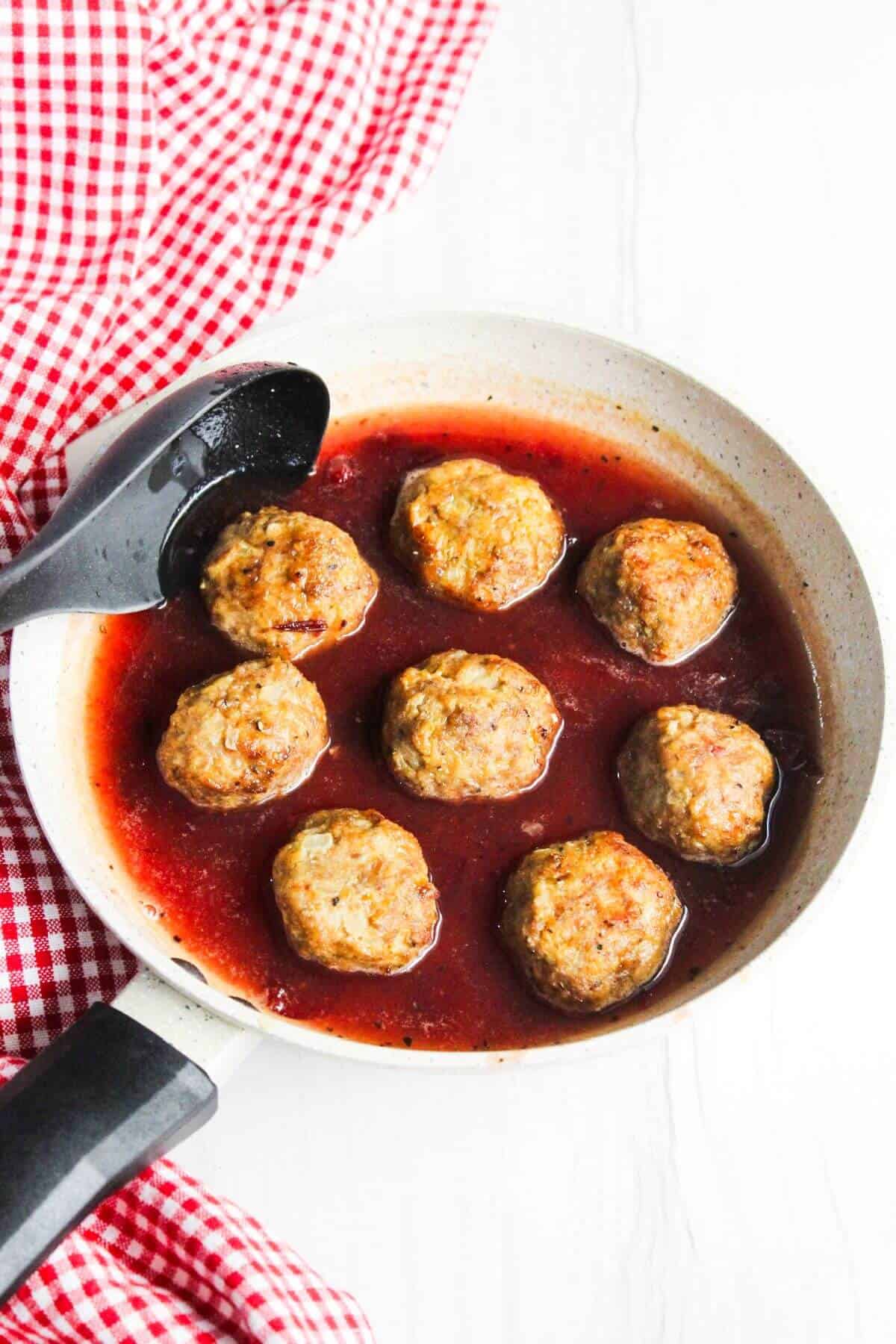 A frying pan with seven ground turkey meatballs simmering in a red sauce, placed beside a red and white checkered cloth. A black spoon rests in the pan.
