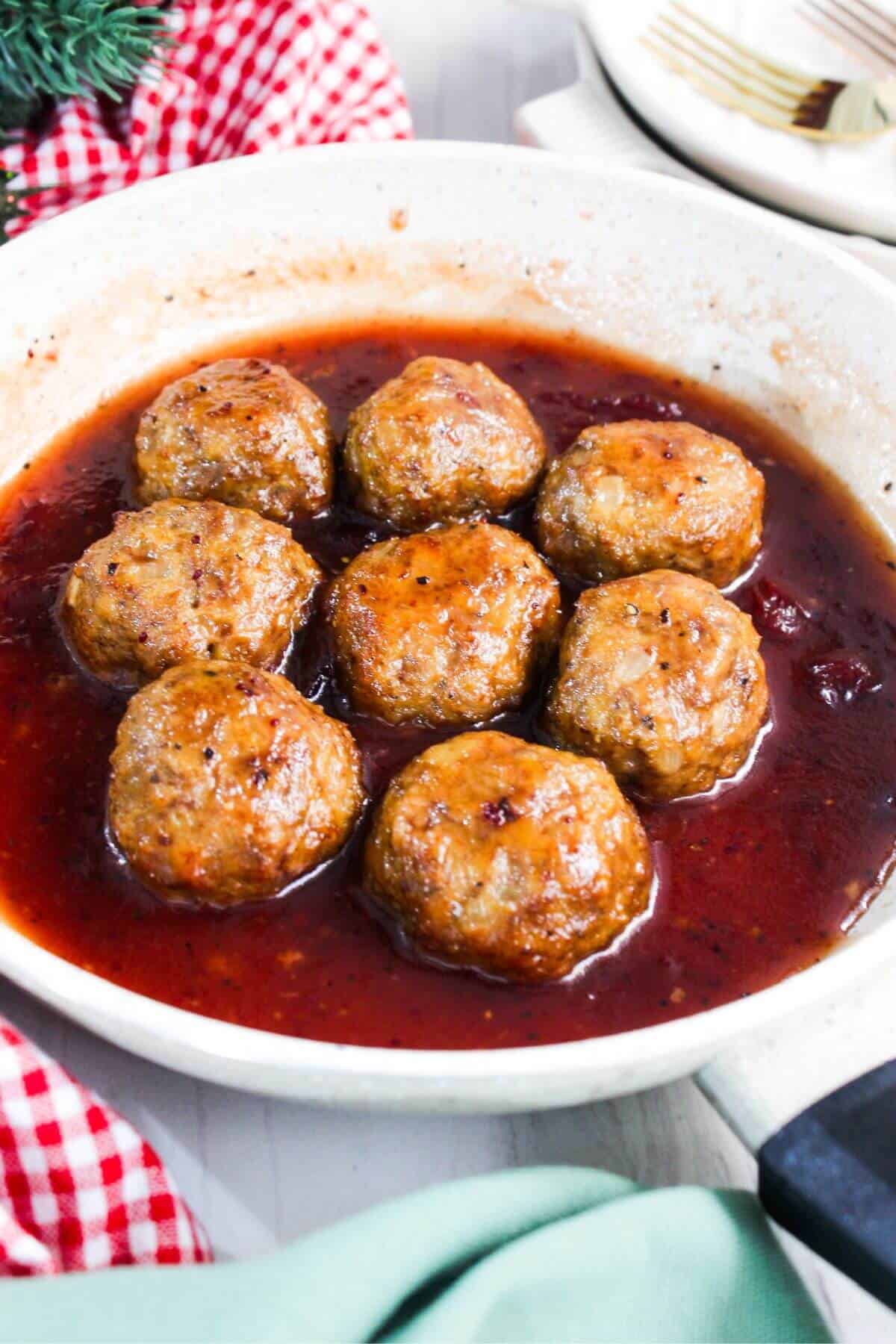 A white skillet contains eight ground turkey meatballs covered in a red sauce. A red and white checkered cloth is partially visible on the left side. Plates and a fork are in the background.
