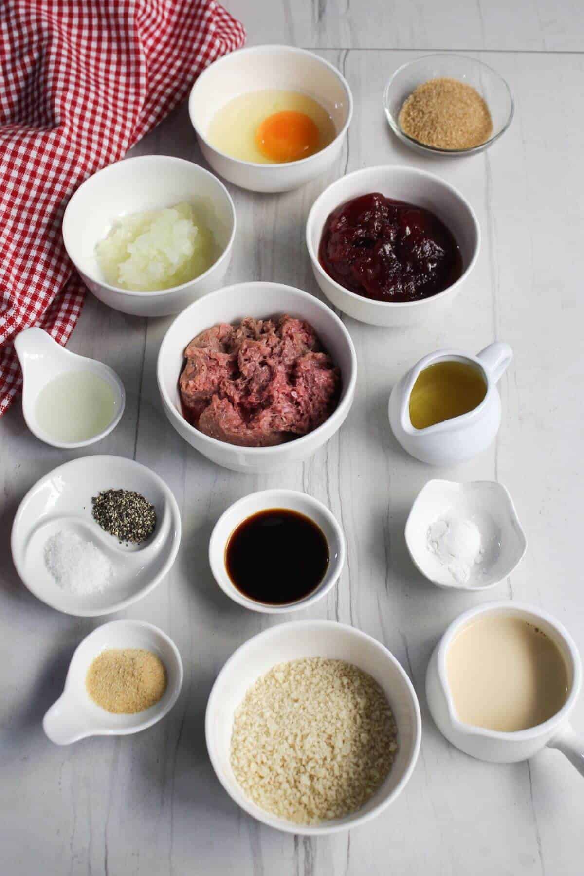 An assortment of ingredients including ground meat, egg, breadcrumbs, spices, sauces, and liquids are arranged in white bowls on a light gray surface next to a red and white checkered cloth.