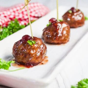 Three glazed ground turkey meatballs garnished with herbs and a red topping, each skewered with a toothpick, served on a rectangular white plate.